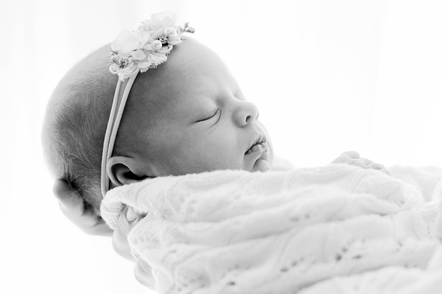 A peaceful sleeping newborn baby wrapped in soft white blanket wearing delicate headband in black and white portrait.