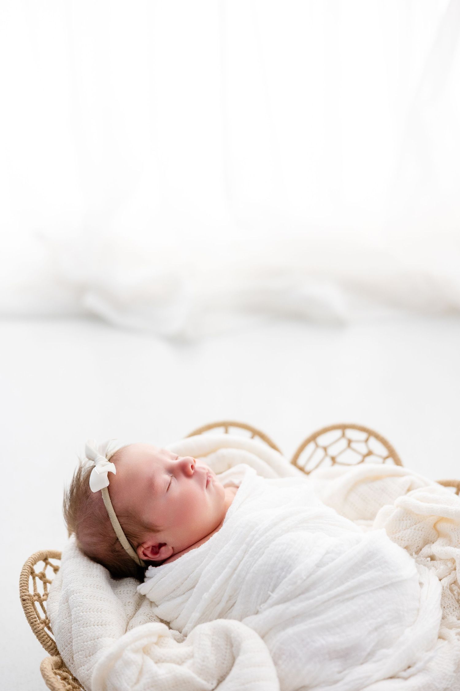 A sleeping newborn baby swaddled in white fabric lies peacefully in a woven basket during a professional photoshoot.