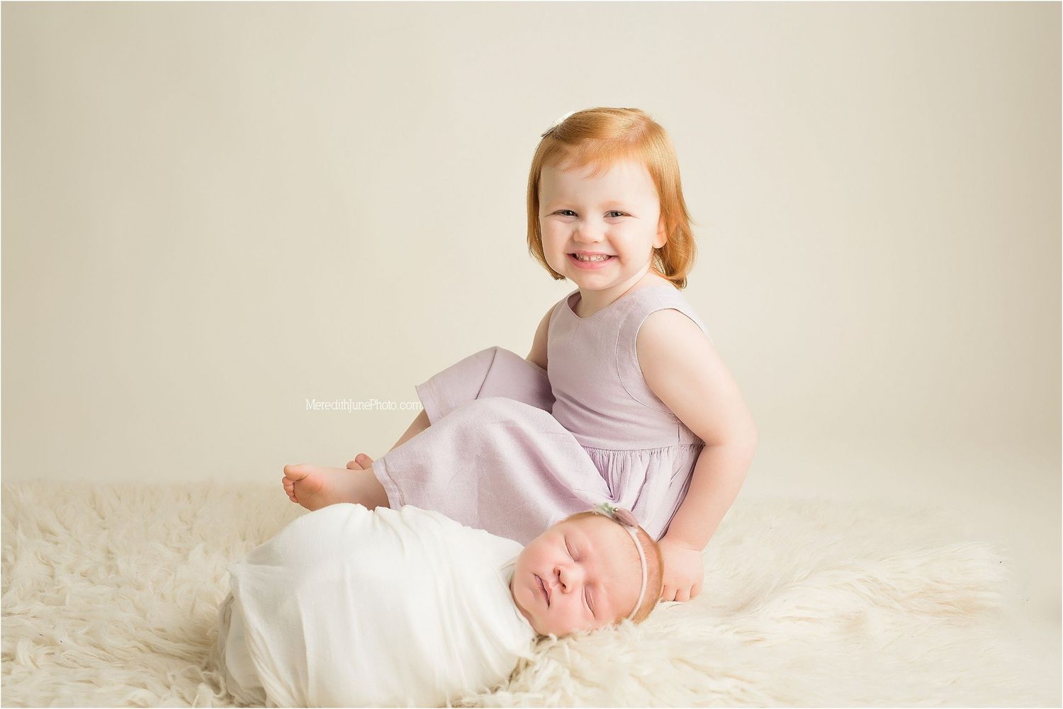 Two young children in light colored clothing sit together on a soft white rug during a portrait session.