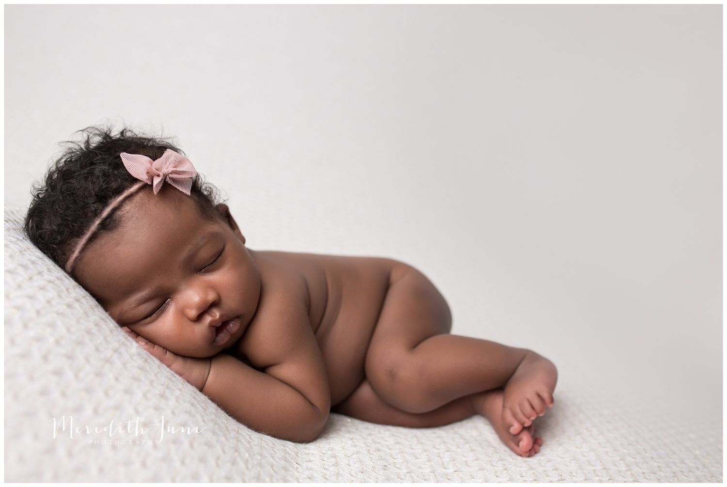 Peaceful sleeping newborn baby with pink bow headband lying on soft white blanket in studio setting.