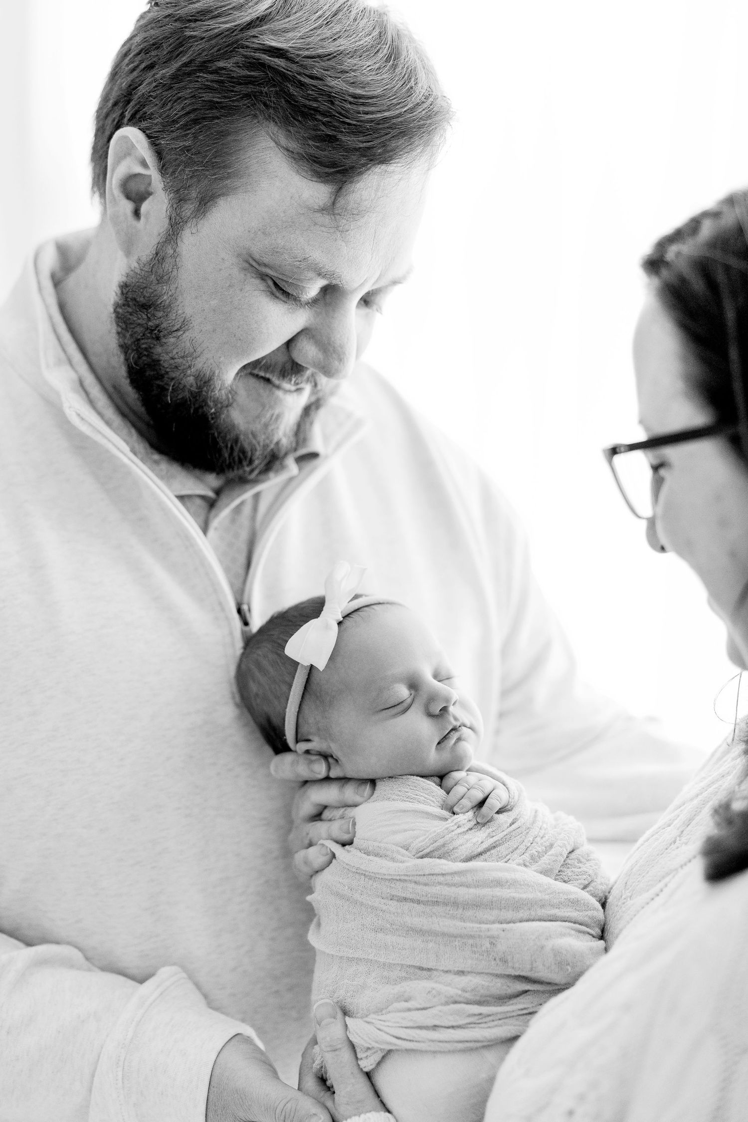 A tender black and white portrait of parents sharing a quiet moment with their sleeping newborn wrapped in blankets.
