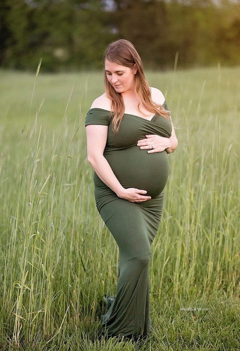 A pregnant woman in a green dress stands thoughtfully in a sunlit field of tall grass during a maternity photoshoot.