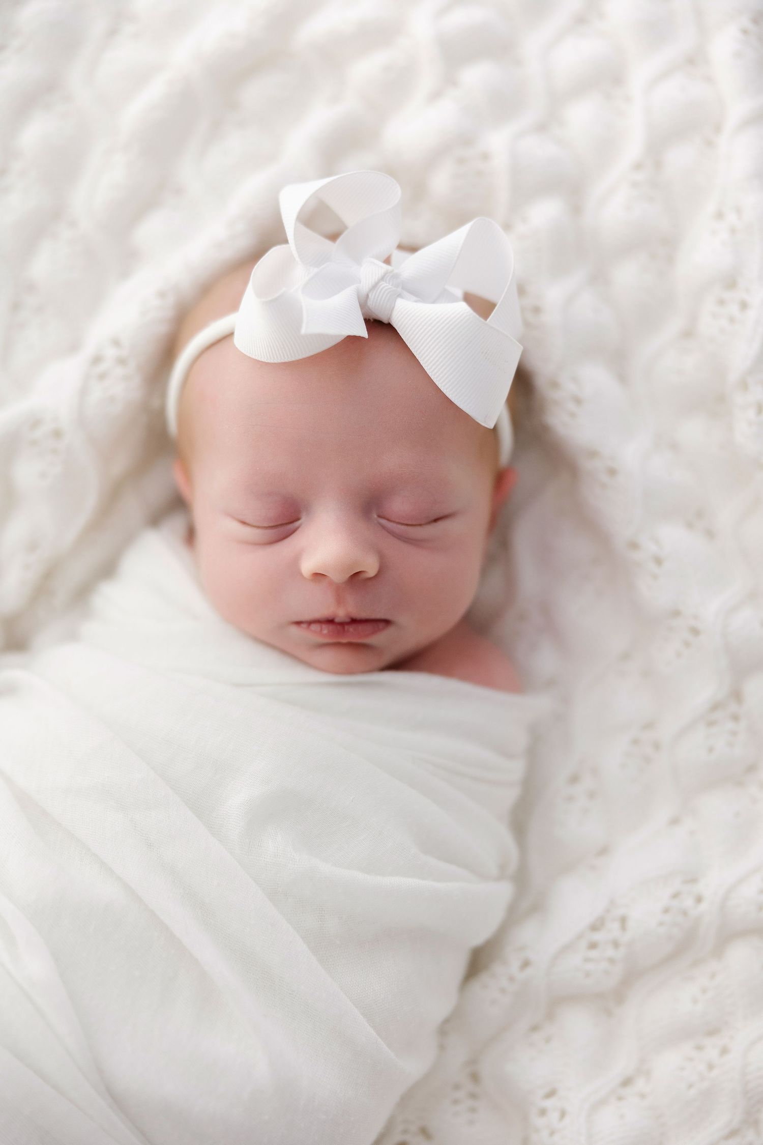 A sleeping newborn swaddled in white fabric with a delicate white bow rests peacefully on textured bedding.