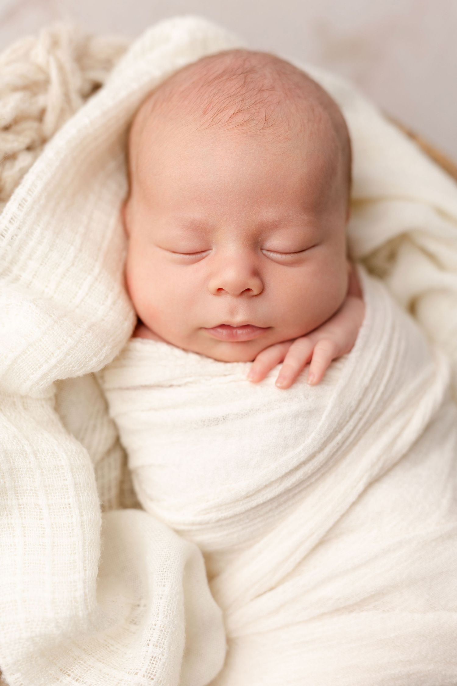 A peaceful sleeping newborn baby swaddled in soft white blankets against a cream-colored background.