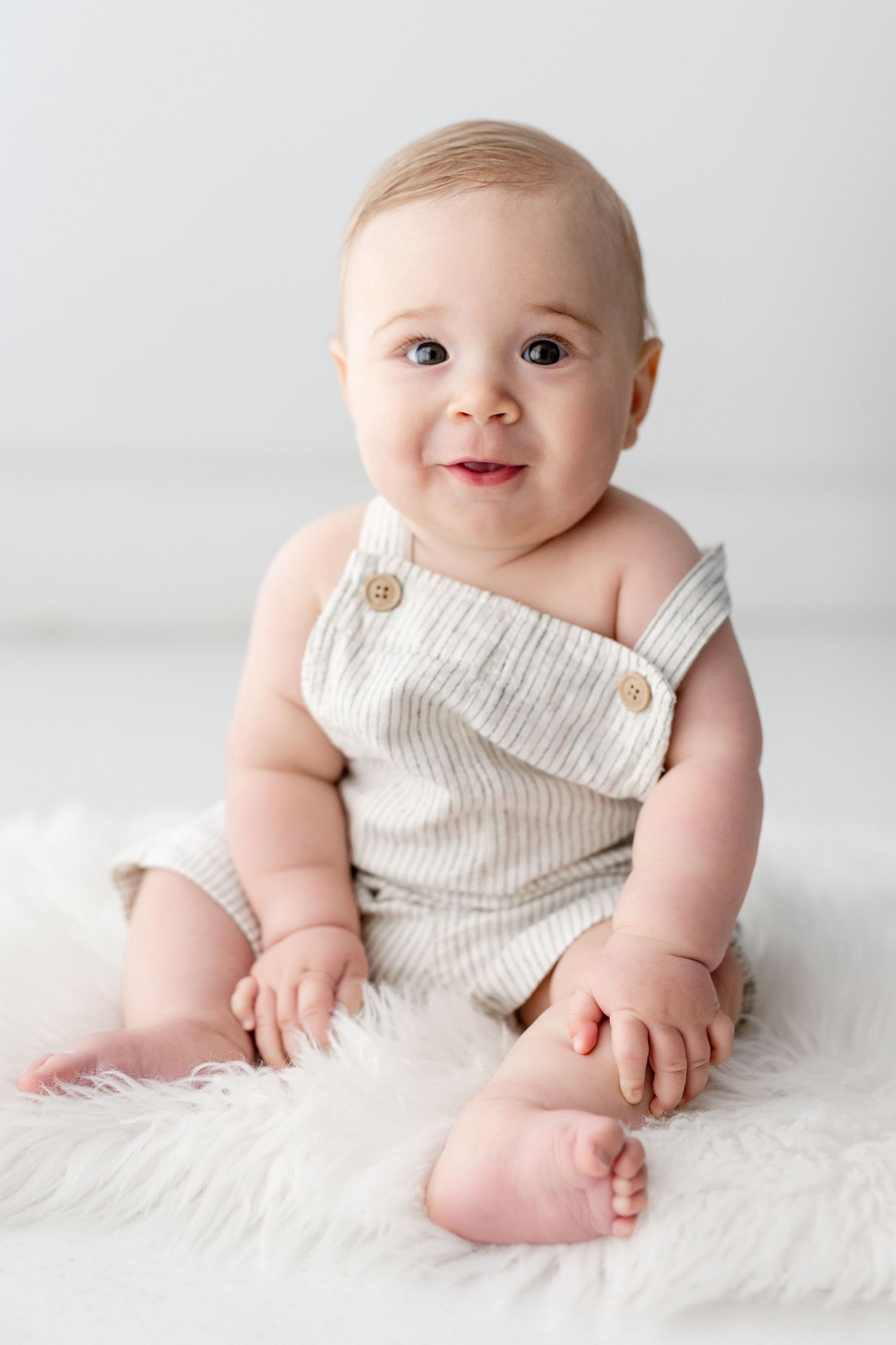 Adorable baby wearing white ribbed romper sits on fluffy white rug in progression of candid poses.