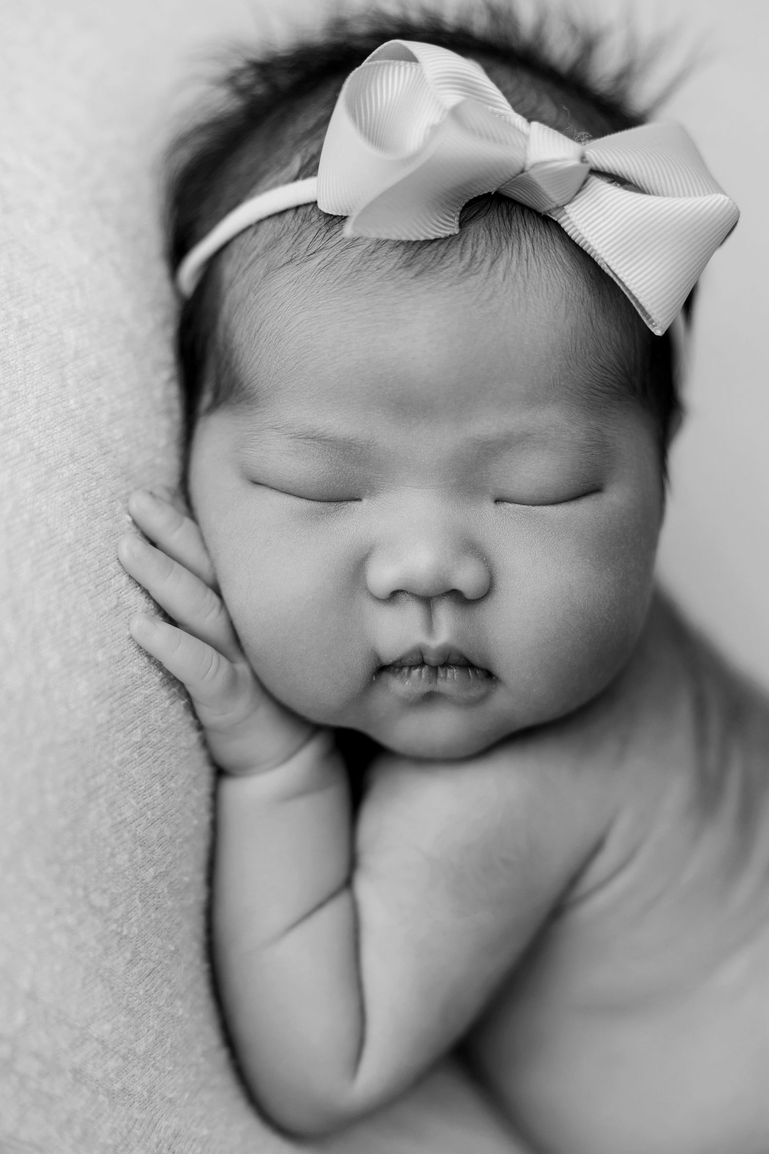 Black and white newborn portrait of a sleeping baby girl wearing a ribbon bow headband, peacefully resting her cheek on her hands.