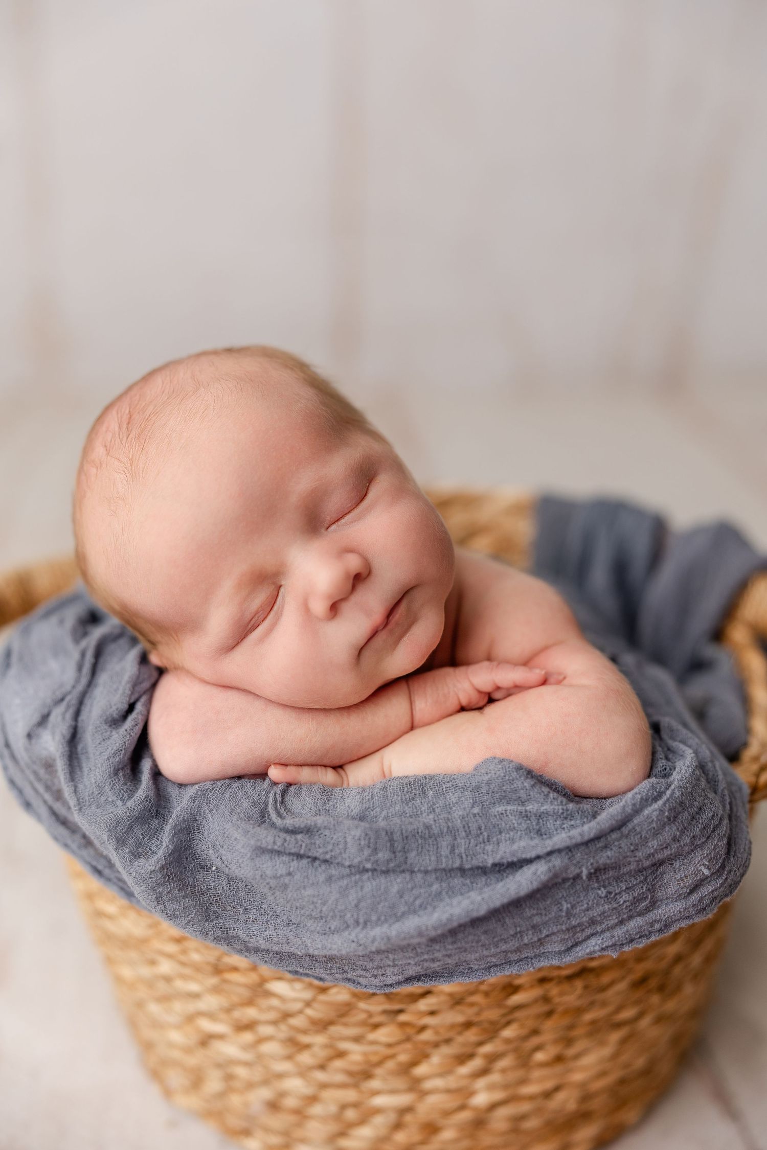 Sleeping newborn baby wrapped in blue cloth nestled in a woven basket for a professional newborn photo session.