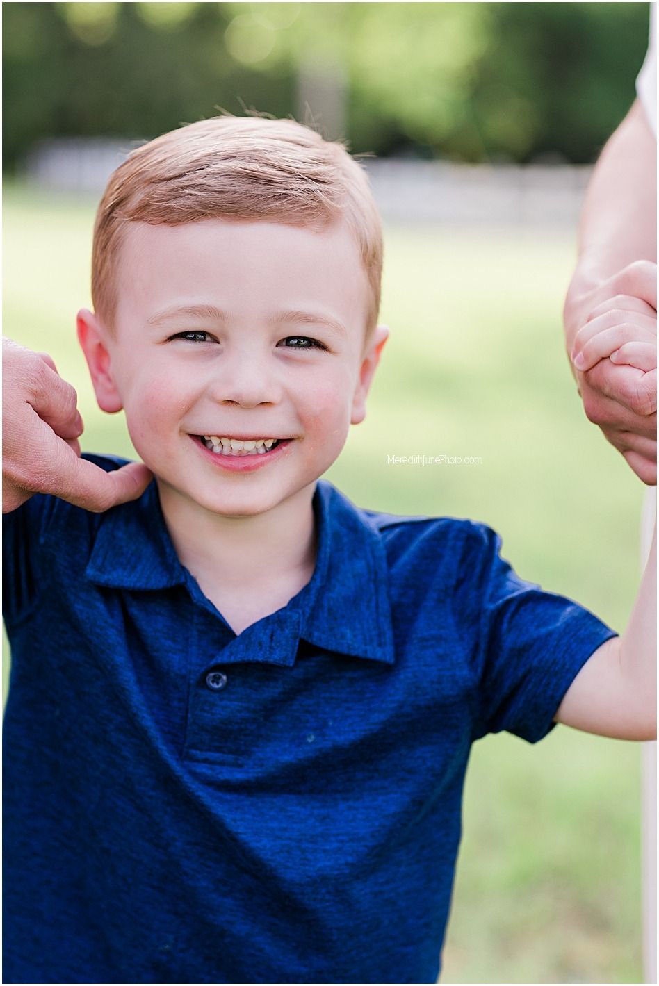 A child in a navy blue polo shirt smiles brightly while playing outdoors on a sunny day.