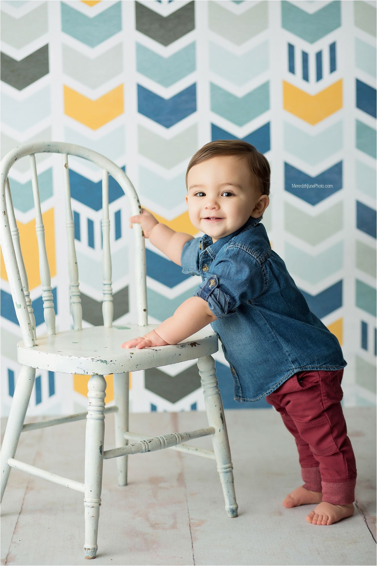 Baby in denim top plays with white vintage chair against geometric patterned background.