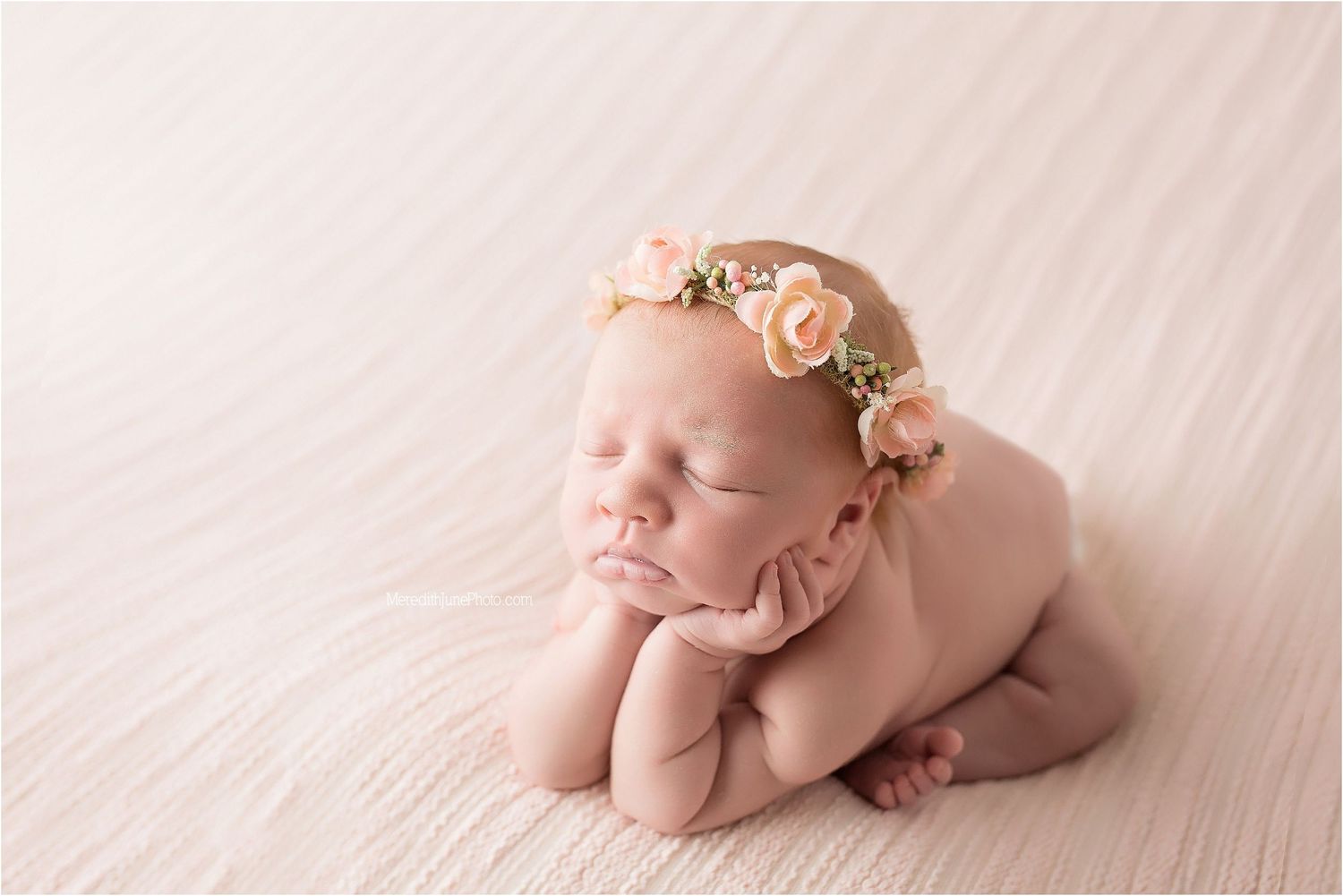 A sleeping newborn in a peach floral headband lies curled up on soft pink fabric in a studio setting.