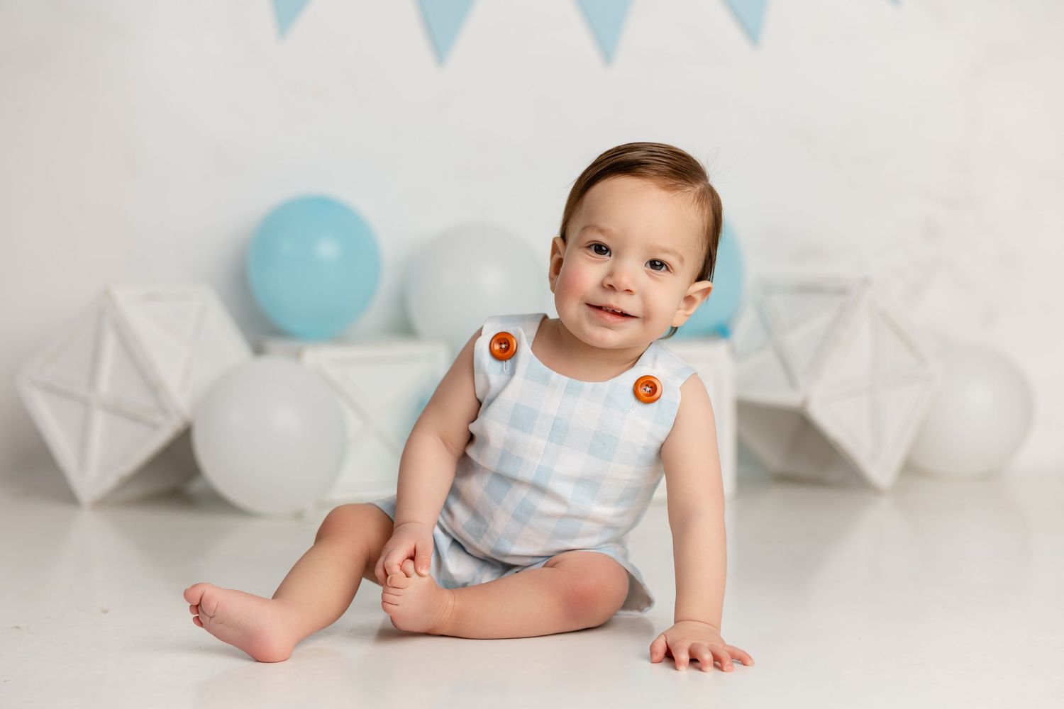 Baby wearing light blue romper sits on white floor with blue balloon and white decorations in background.