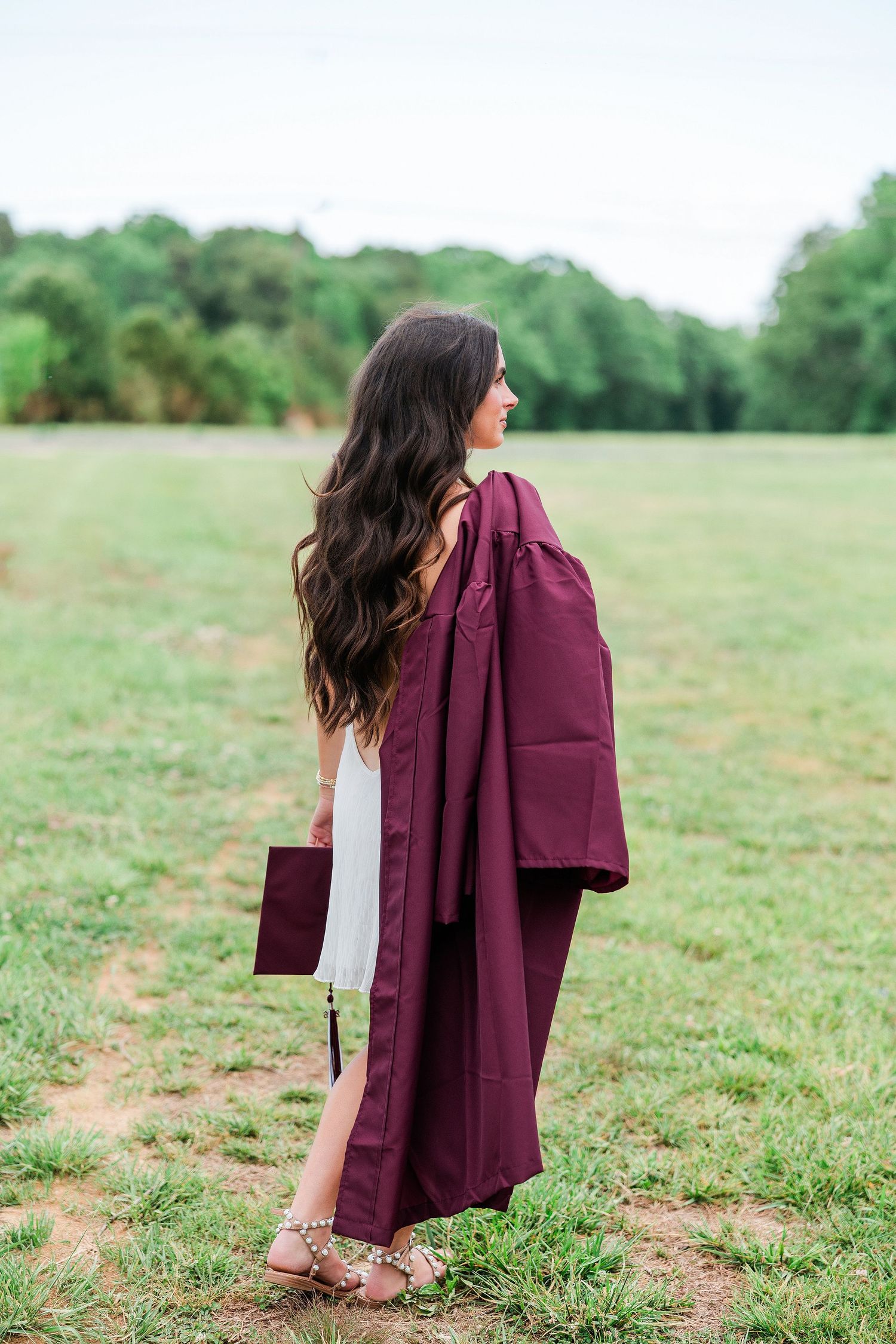 Young woman holding maroon graduation gown and cap in a green field, wearing a white dress and heeled sandals.