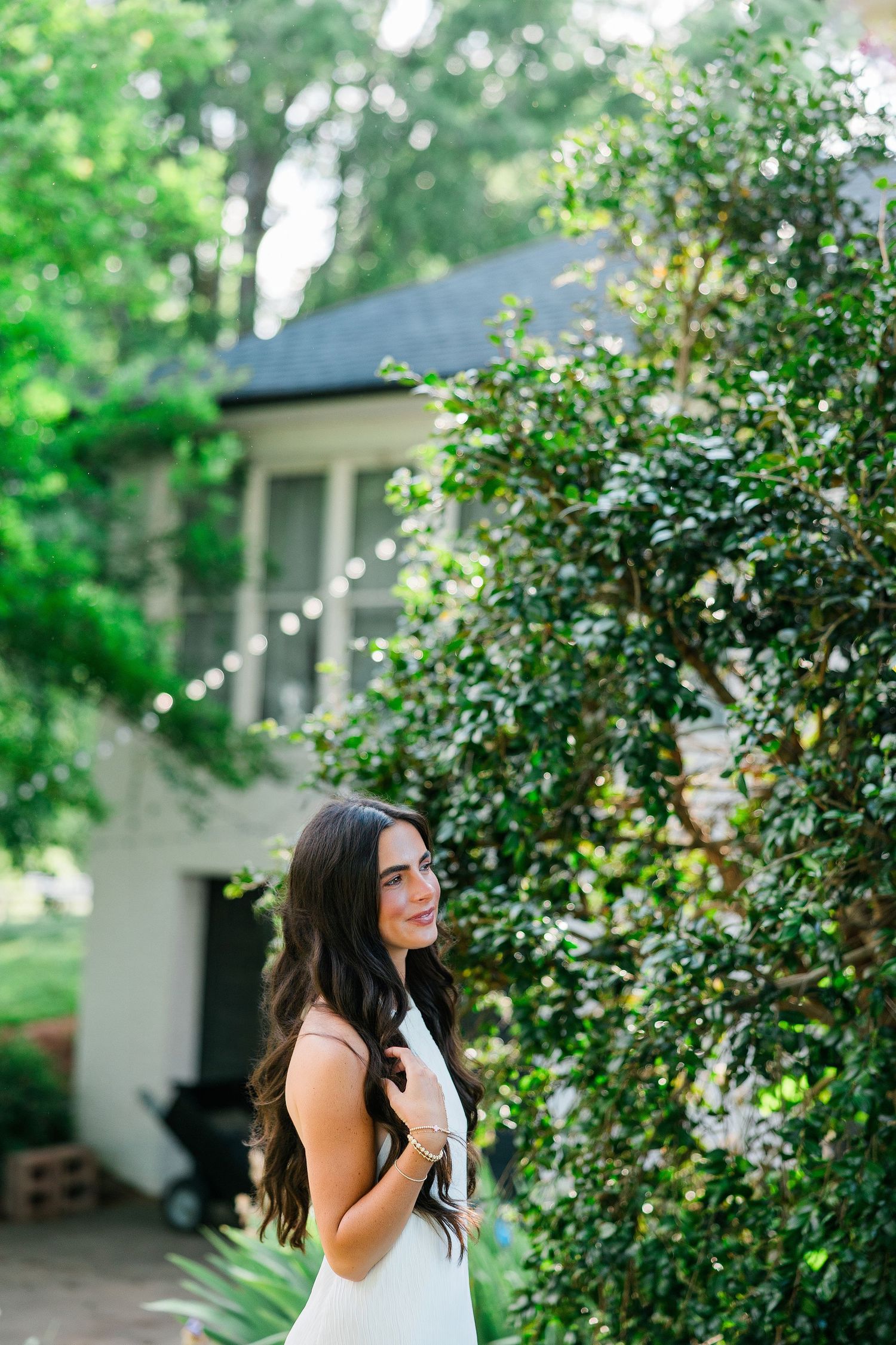 Brunette woman in white dress smiling beside lush green ivy-covered wall near a white house.