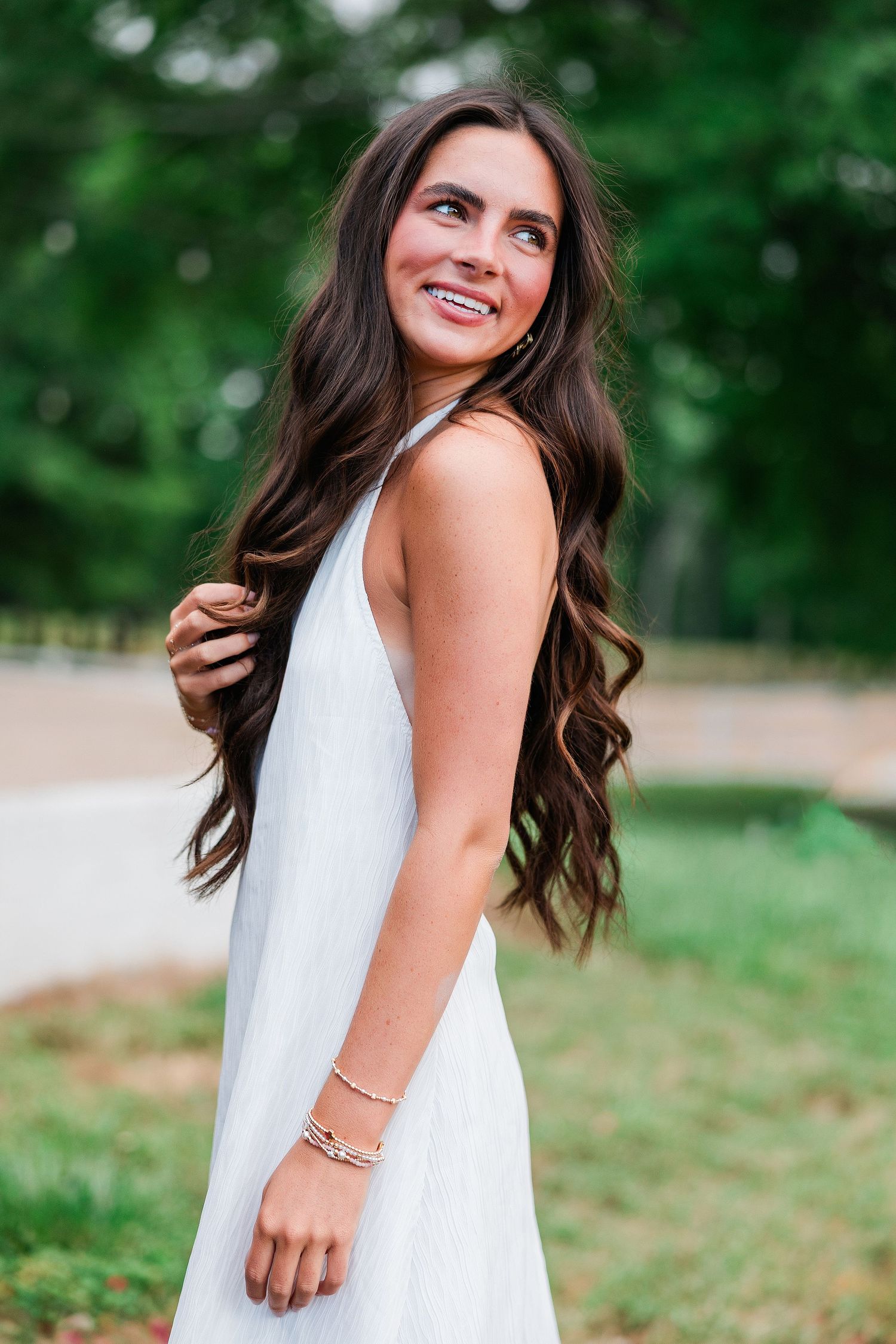 Smiling brunette woman in white dress with long wavy hair posing outdoors in a lush green park.
