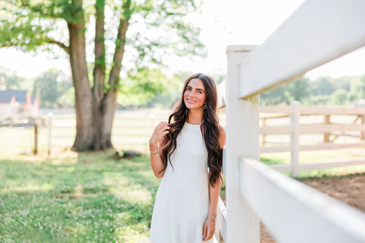 Young woman in white dress smiling outdoors near white fence with lush green trees in sunny farm setting.