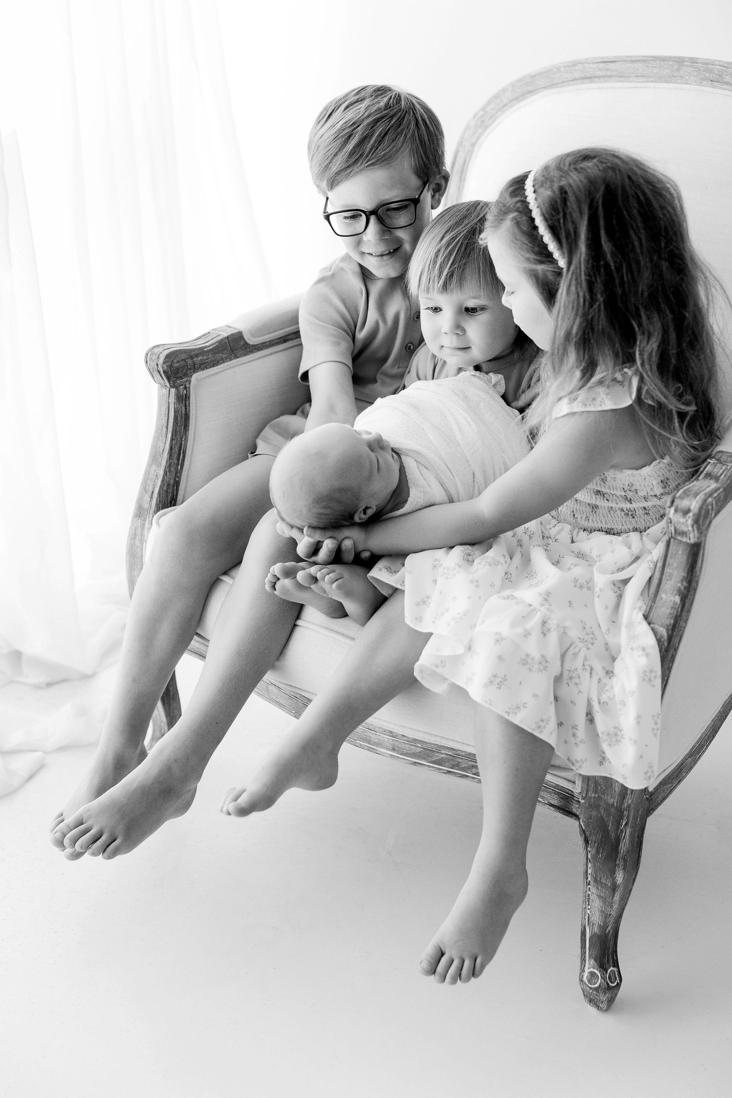Black and white photo of three children sitting in a vintage chair, lovingly holding a newborn baby.