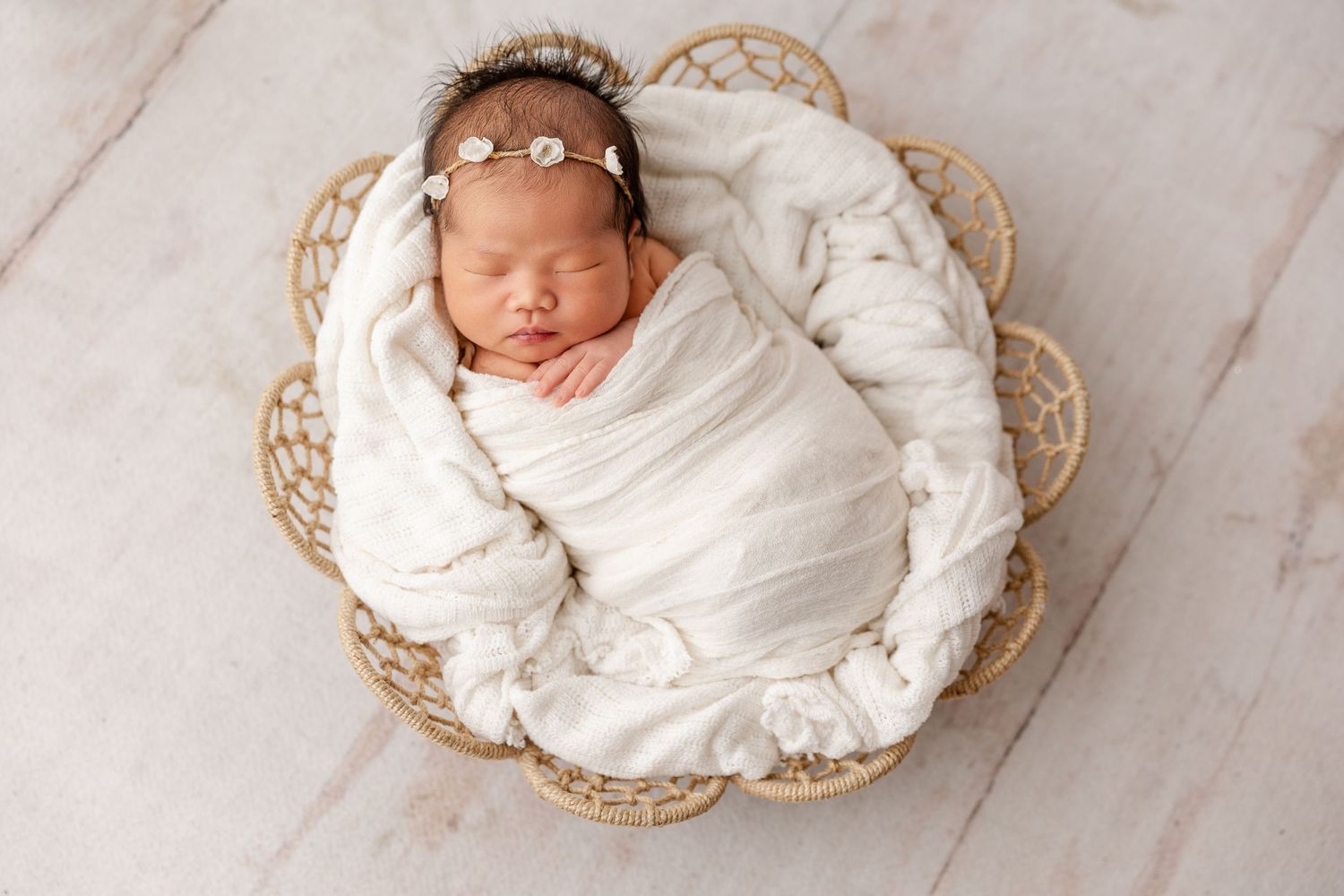 Newborn baby wrapped in white swaddle sleeping in wicker basket with floral headband.