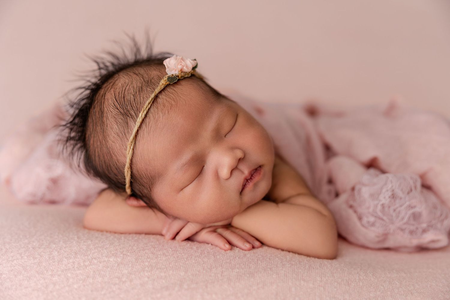 Newborn baby girl sleeping peacefully on pink blanket, wearing floral headband, in professional newborn photography pose.