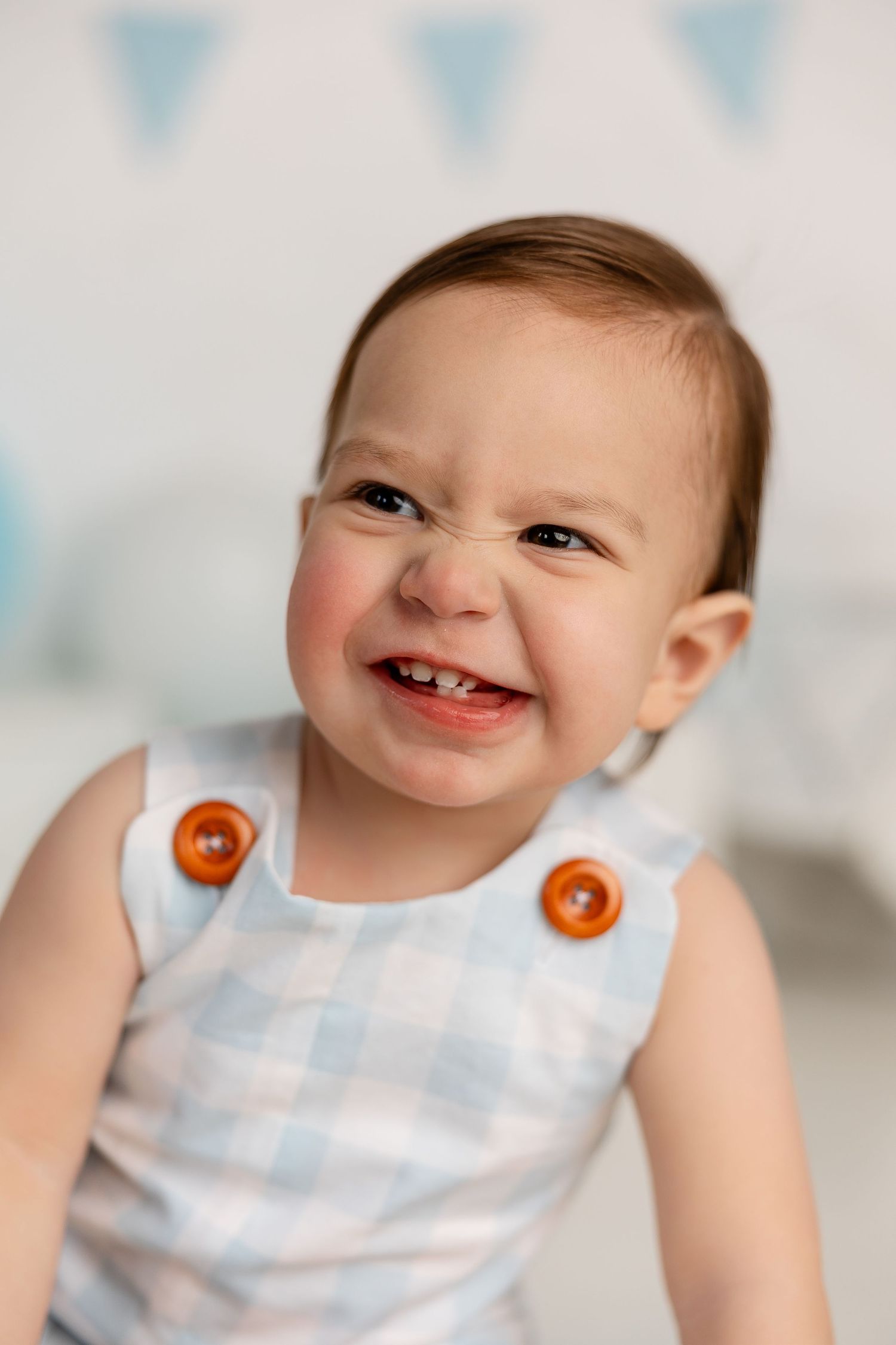 Baby wearing blue and white plaid romper with orange buttons smiles joyfully against light backdrop with blue bunting.