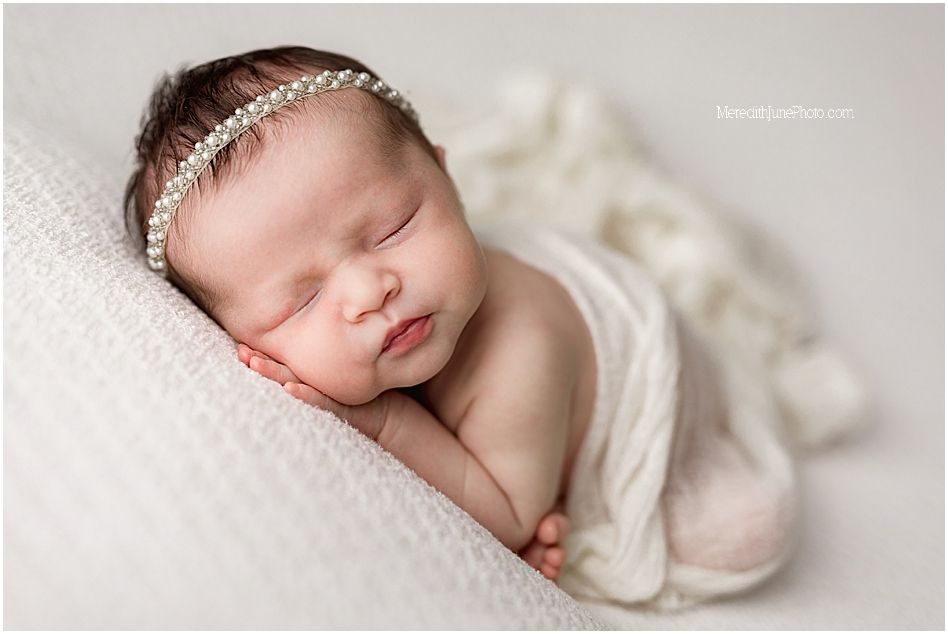 Sleeping newborn wrapped in soft white fabric wearing delicate rhinestone headband on textured white backdrop.