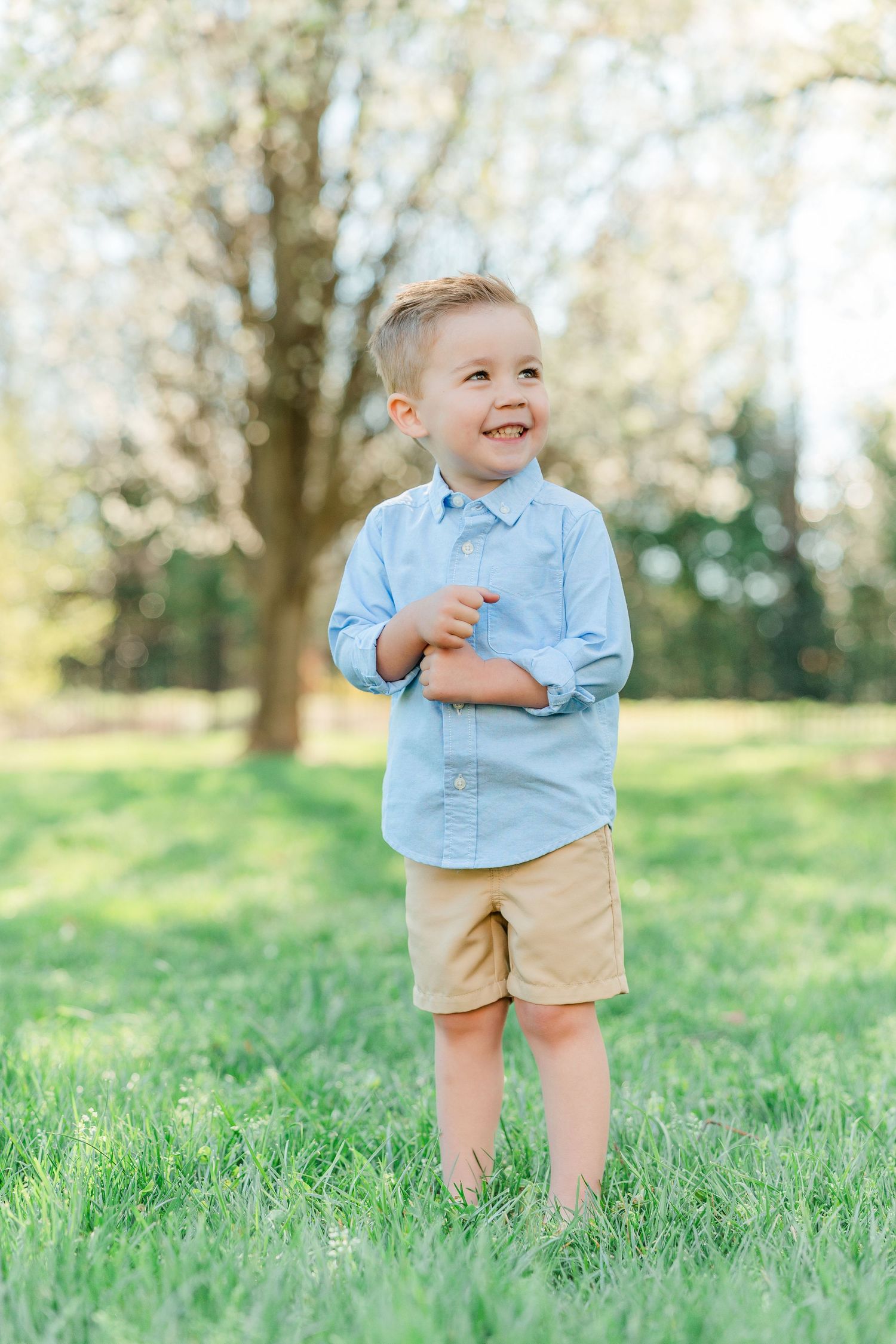 A toddler in a light blue shirt and khaki shorts plays on a grassy hill during springtime.