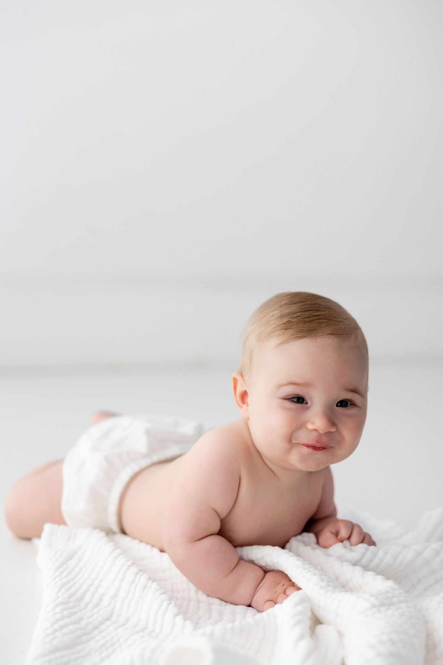 Adorable baby in white outfit lying on cozy white blanket during professional photo session.