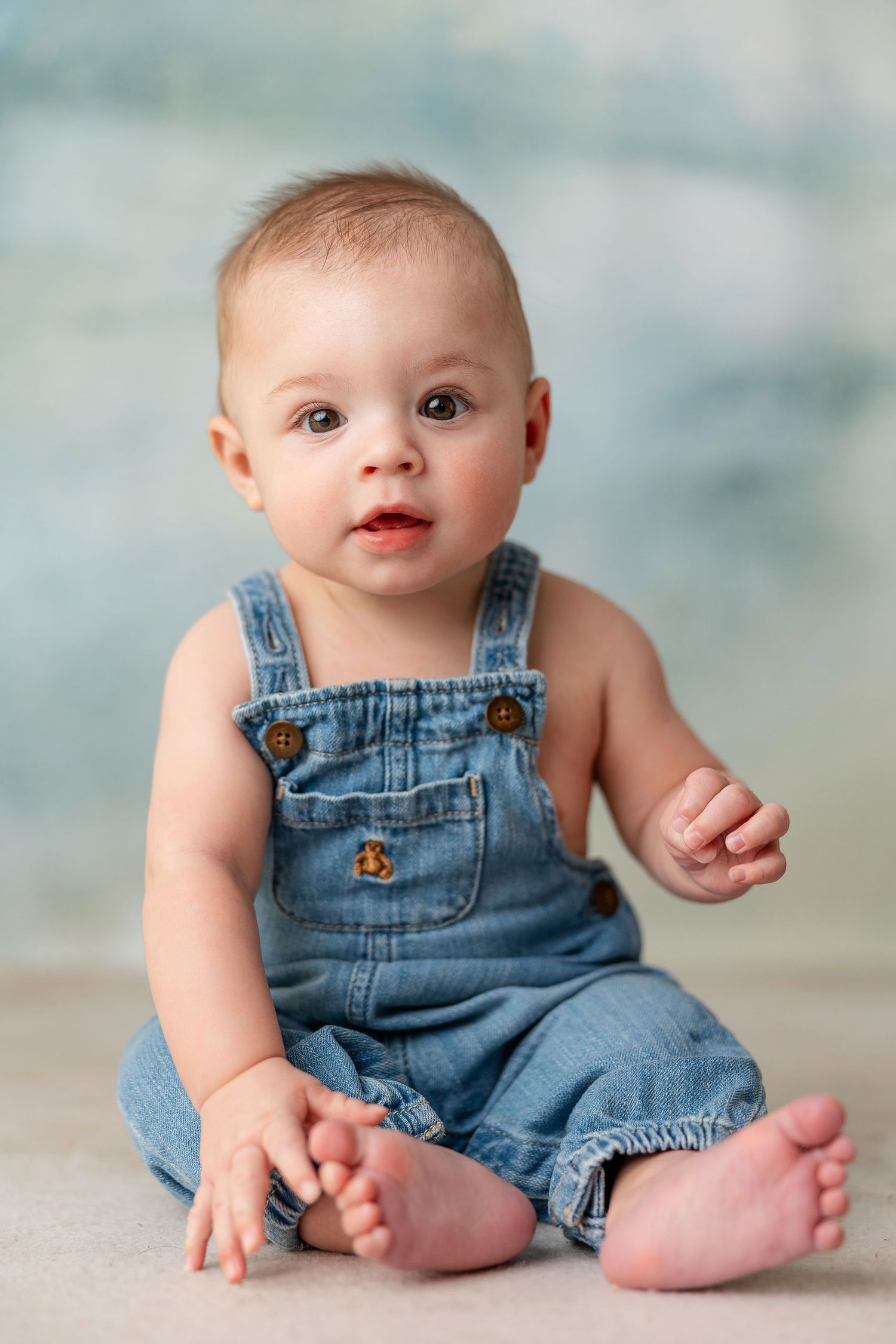 Baby in denim overalls sits and poses against soft blue backdrop during portrait session.