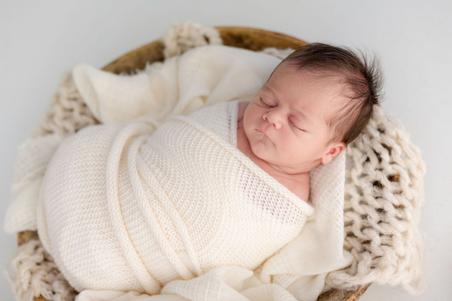 Sleeping newborn baby swaddled in cream knit wrap, nestled in a wicker basket with soft blanket.