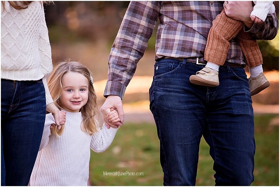 A family holds hands while walking together outdoors, wearing cozy fall clothing in plaid and knit sweaters.