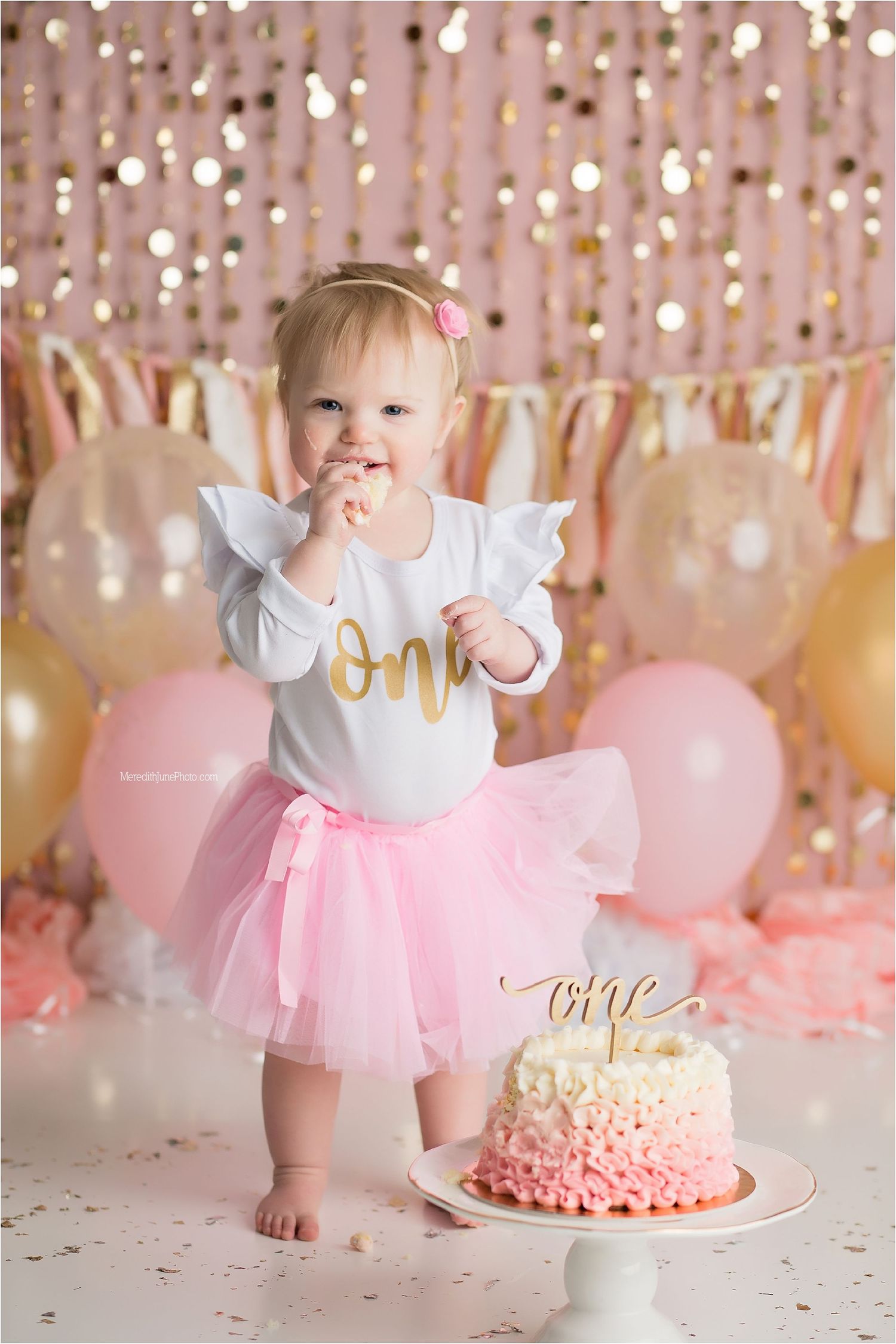 First birthday celebration setup with pink tulle tutu, ombre cake, and sparkly gold backdrop with balloons.