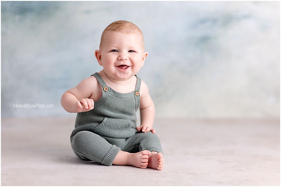 A happy baby wearing a sage green knit romper sits on a light backdrop with a joyful expression.