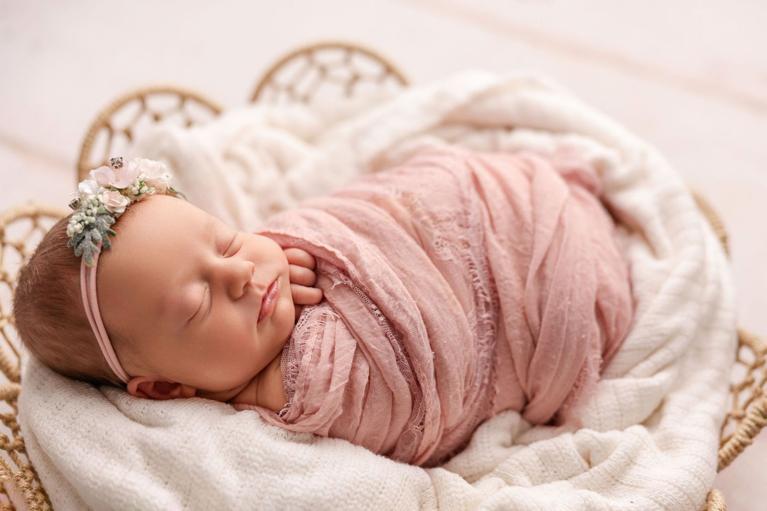 A sleeping newborn wrapped in soft pink swaddle blanket rests peacefully in a woven basket with white lining.