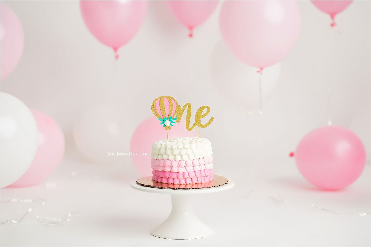 Pink ombre birthday cake with gold 'One' topper on white stand surrounded by pink balloons.