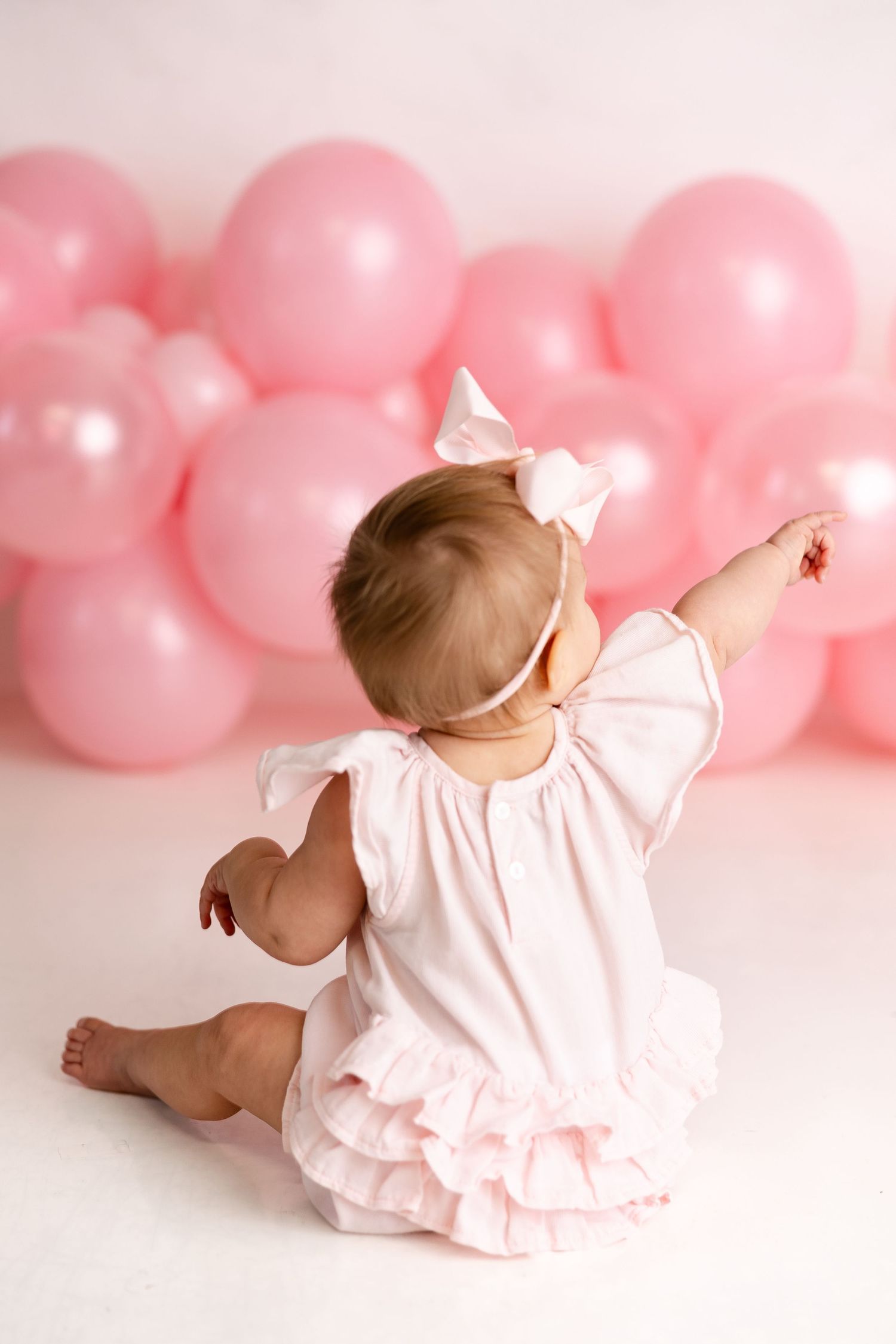 Baby in pink dress sits against pink balloon backdrop in sweet studio photography session.