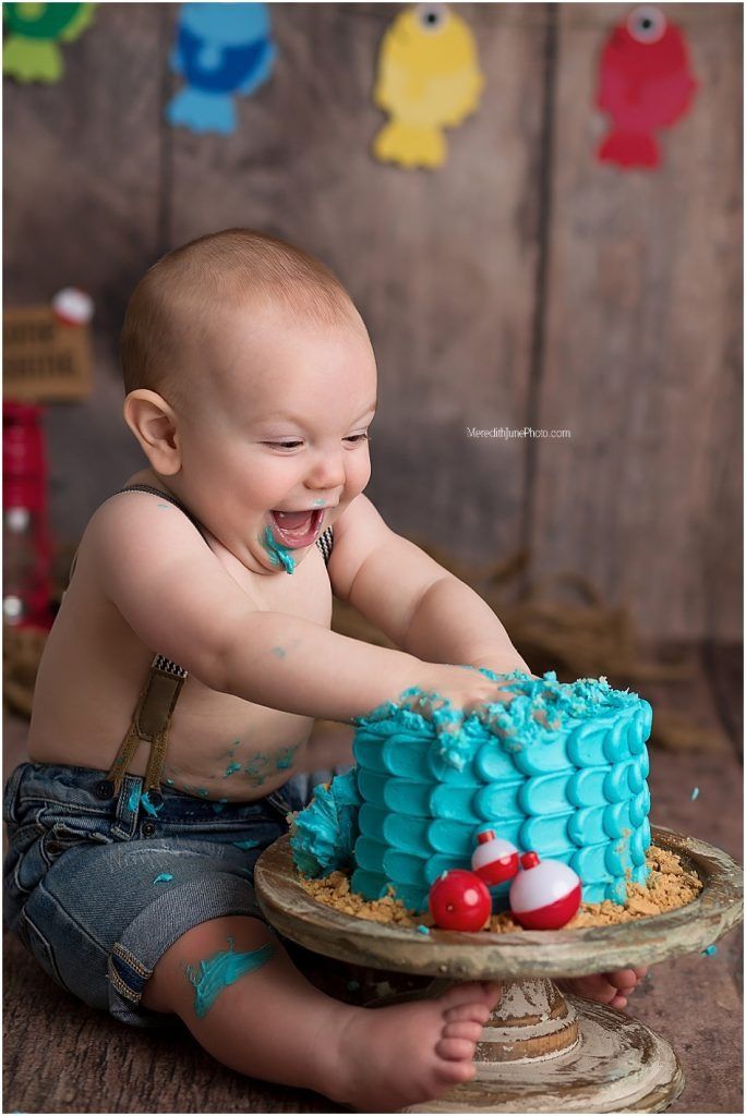 A baby in denim enjoys smashing and playing with a bright turquoise birthday cake during a cake smash photo session.