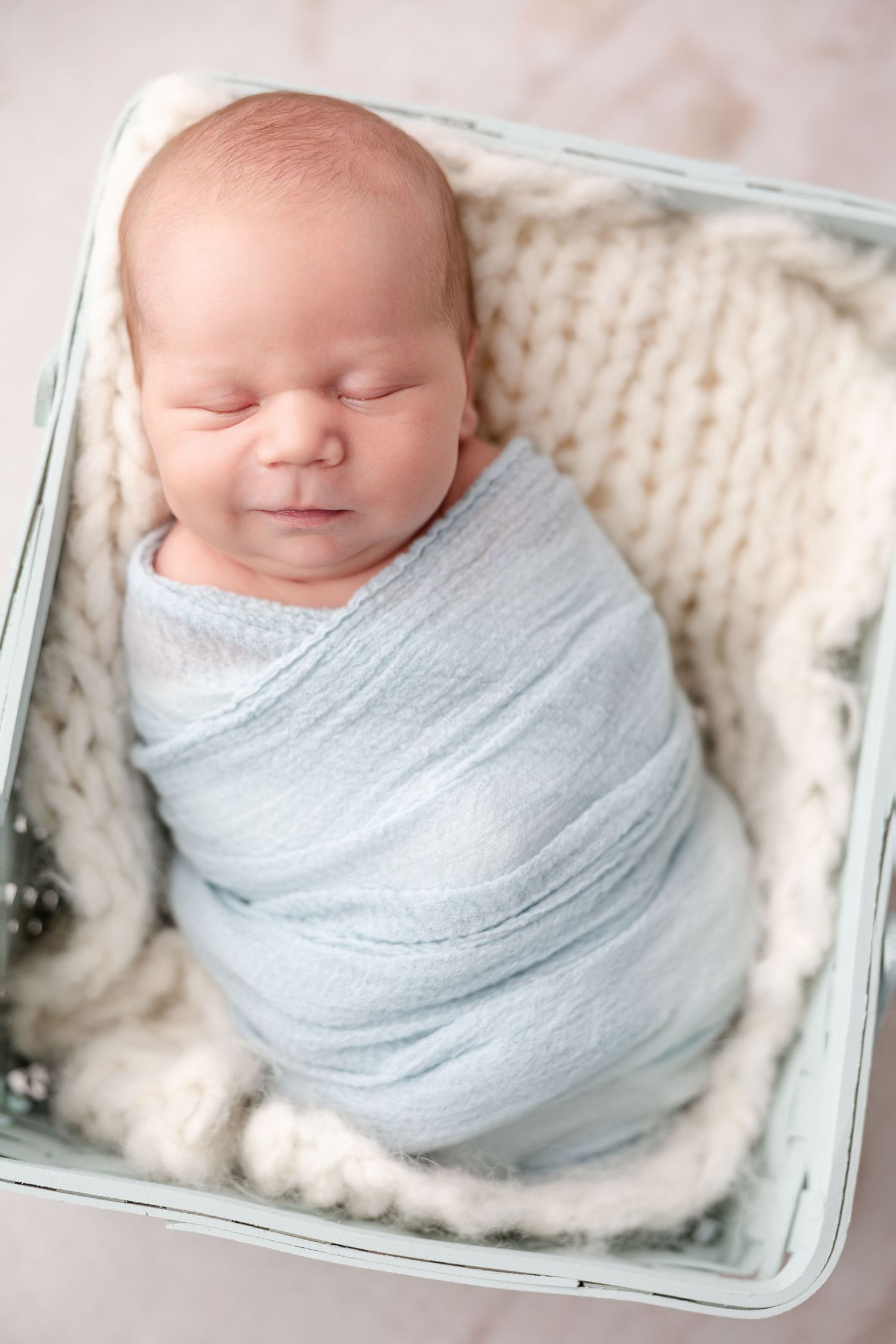 A sleeping newborn baby swaddled in soft blue fabric lying in a white wooden crate on a knitted blanket.