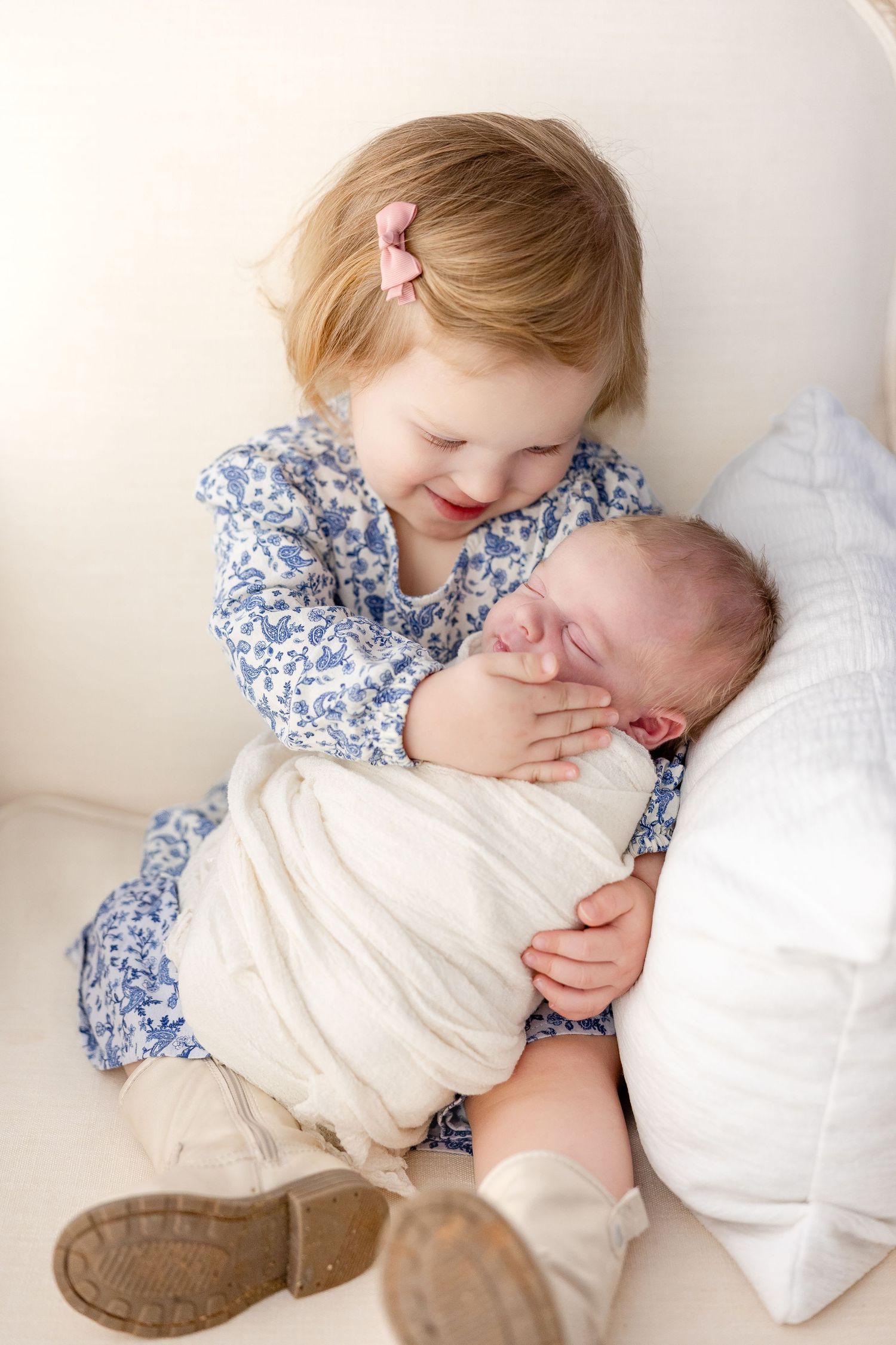 A child in a blue floral dress gently embraces and kisses a swaddled newborn on white bedding.