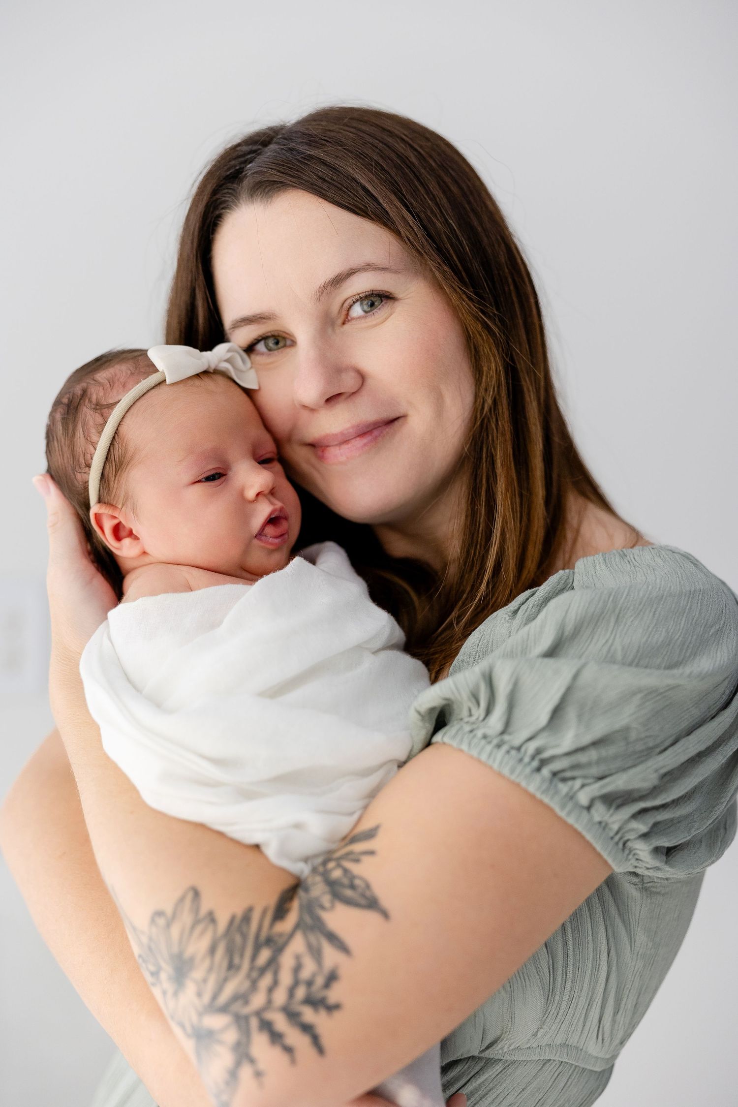 Mother in grey shirt with tree tattoo holds swaddled newborn against white background.