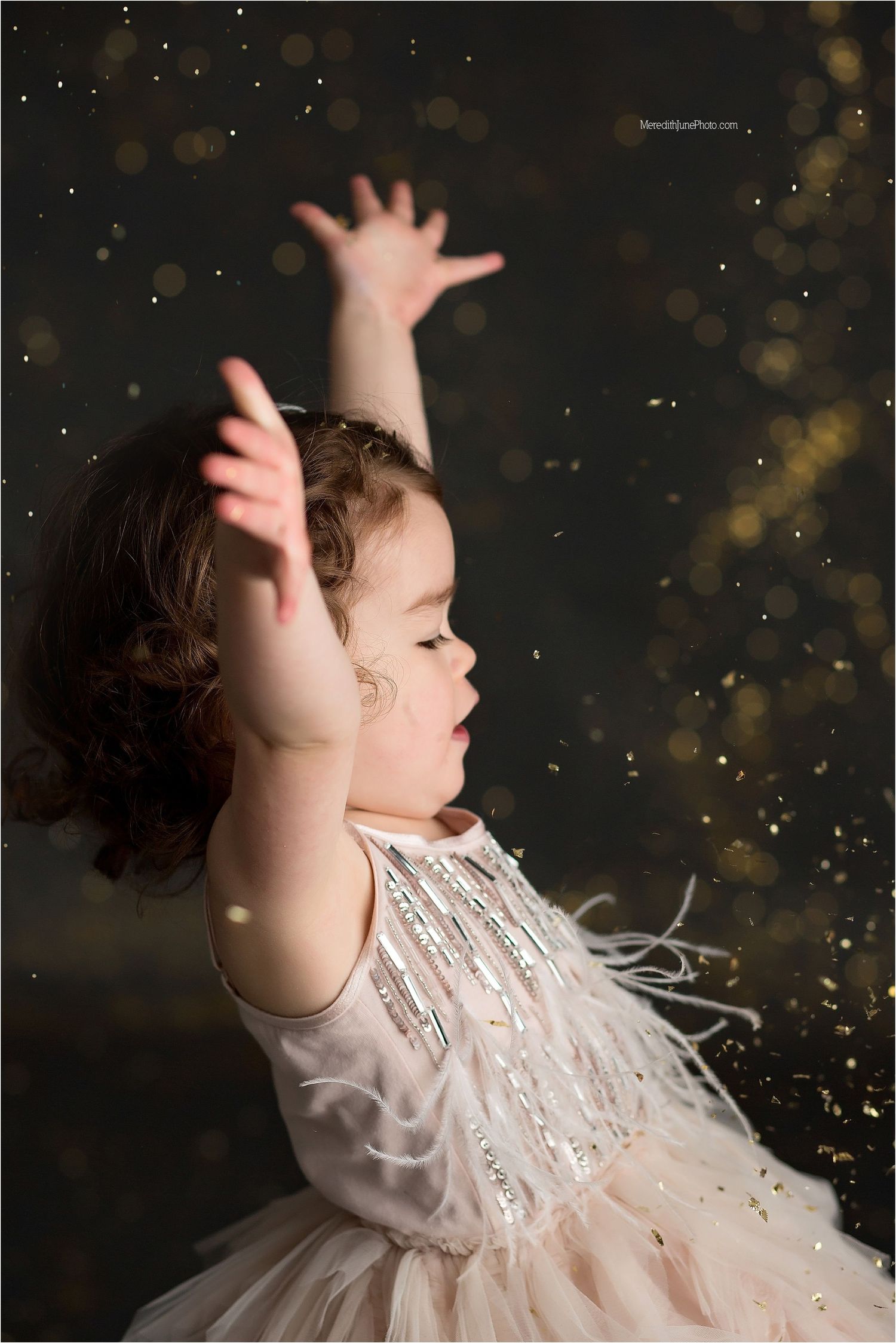 A child in a white lace dress twirls joyfully with raised arms as glitter sparkles in the dark background.