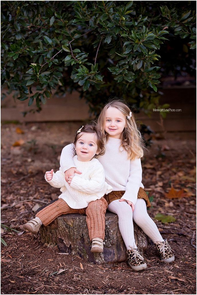 Two children in white sweaters and brown boots sit together on a log surrounded by fallen leaves in an outdoor setting.