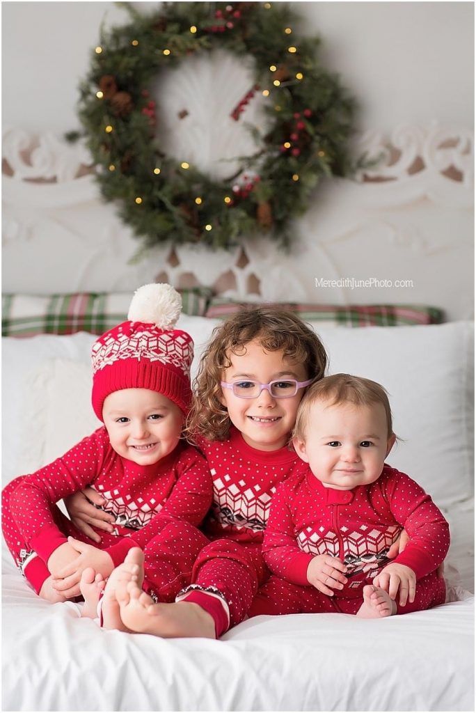 Three children in matching red holiday sweaters sit together on white bedding beneath a Christmas wreath.