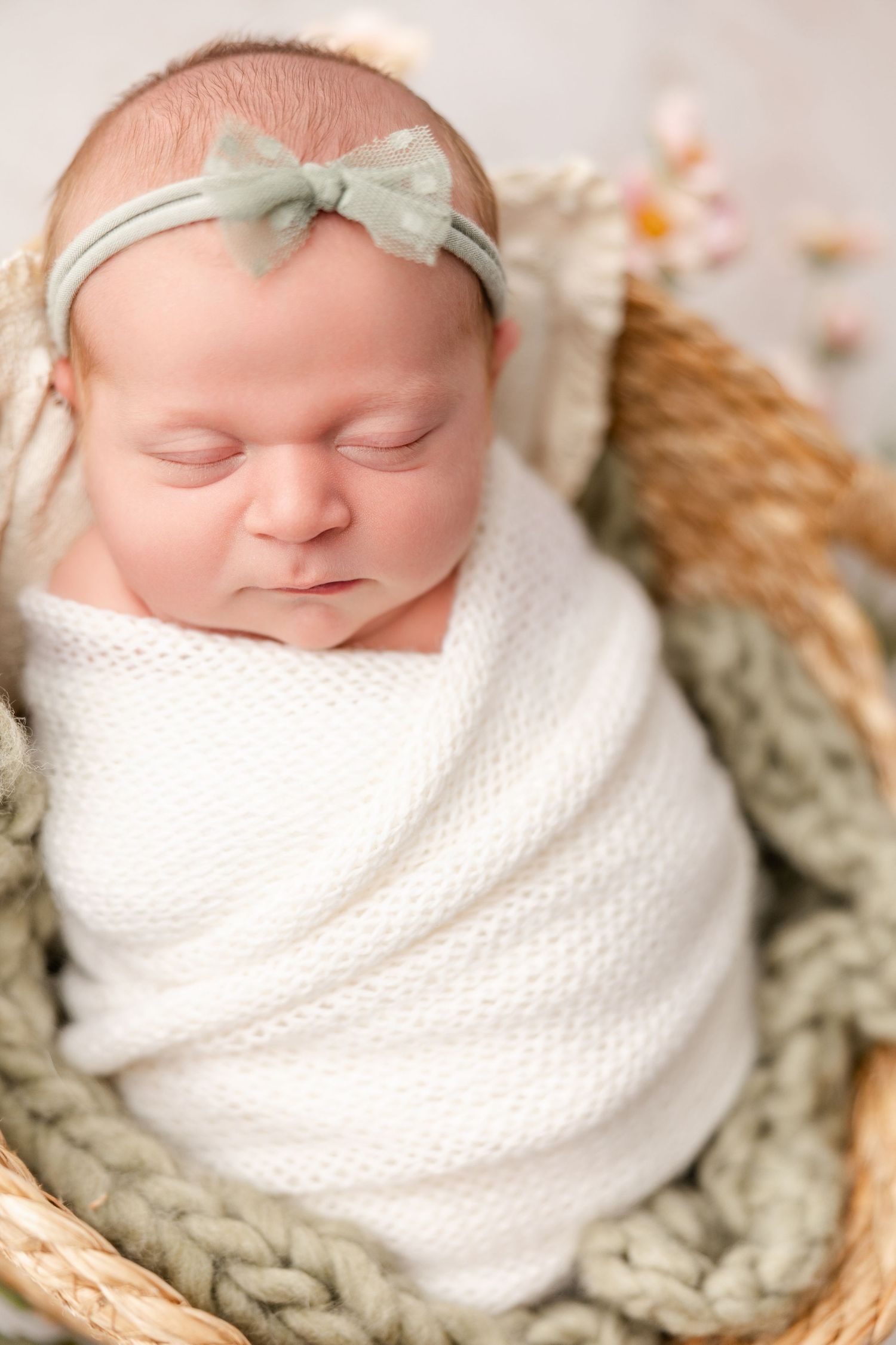 A sleeping newborn baby wrapped in white blanket rests peacefully in a cozy knitted basket.