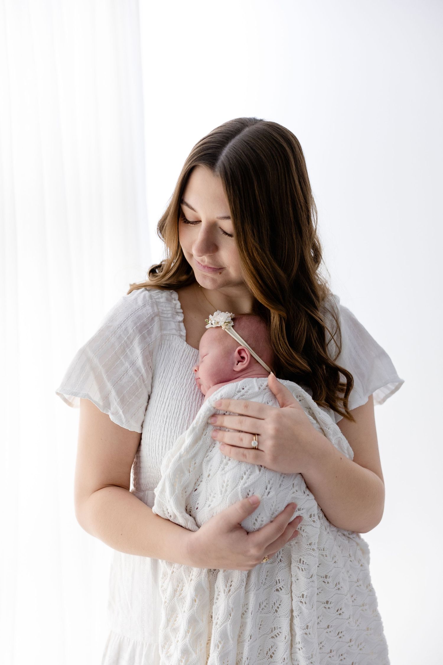 A woman in a white eyelet dress holds a sleeping newborn baby wrapped in white against a light background.