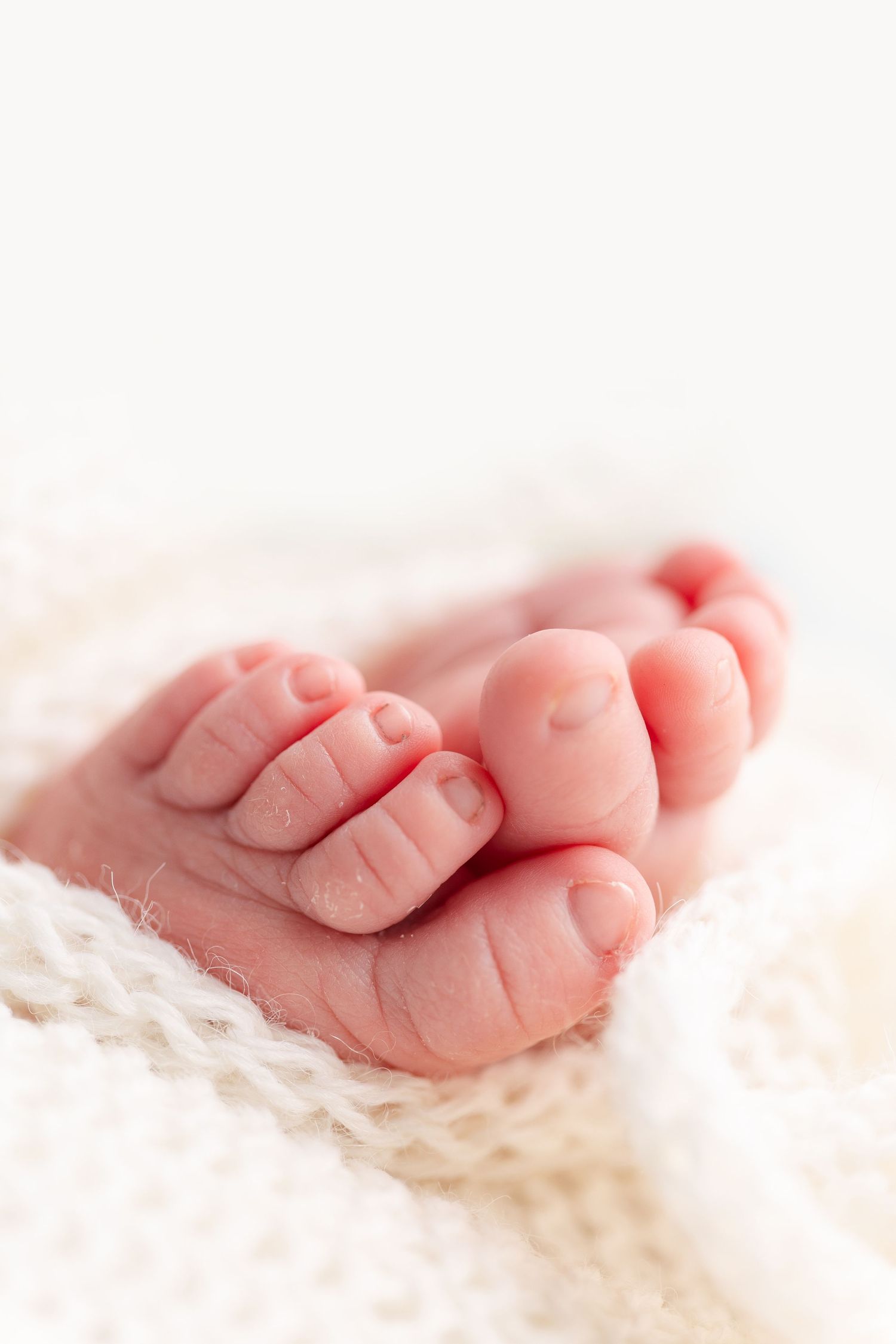 Close-up photo of tiny newborn baby feet resting on soft white textured fabric with gentle pink lighting.