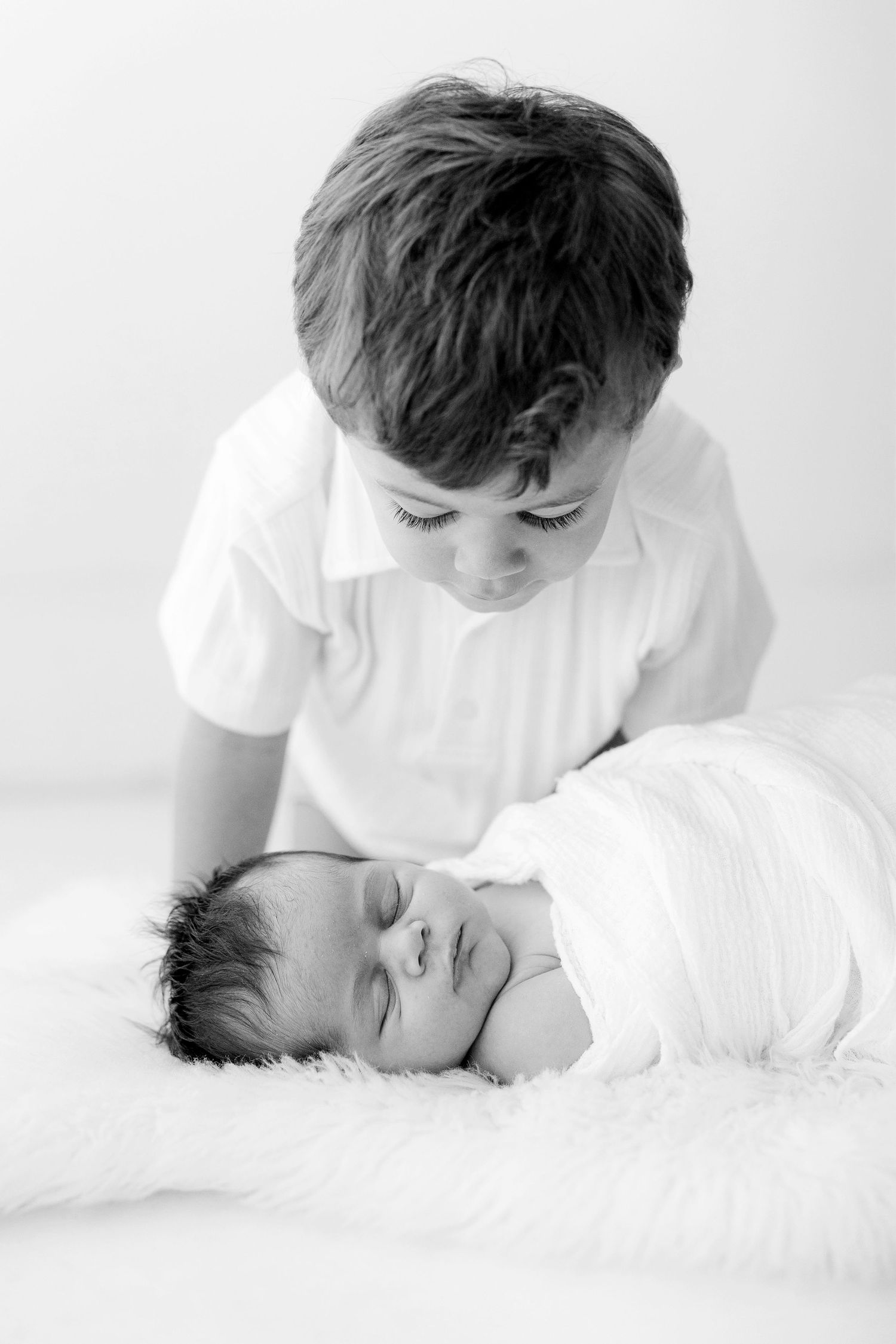 Black and white photo of an older boy tenderly gazing at his sleeping newborn sibling wrapped in white.