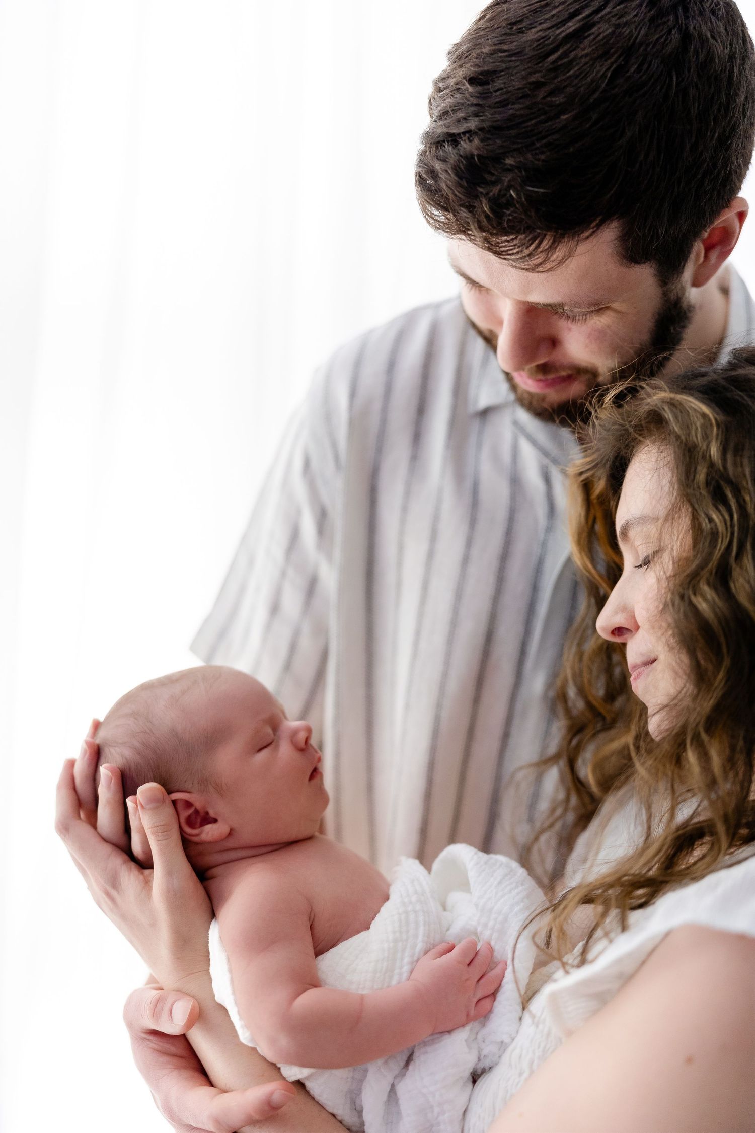 A couple lovingly gazes at their newborn baby wrapped in white, standing together near a bright window.