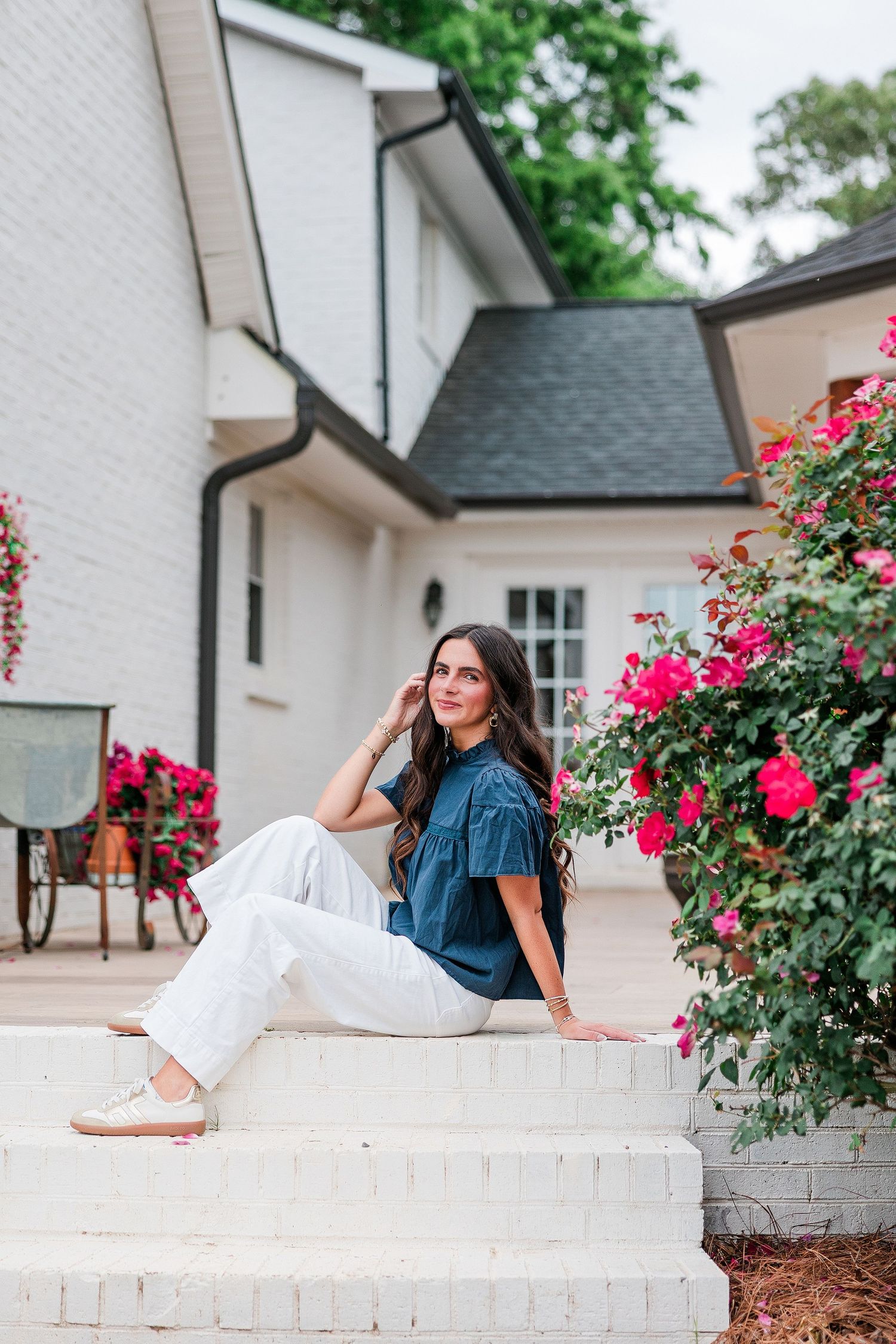 Woman in blue top and white pants sitting on white brick steps near blooming red roses outside a white house.