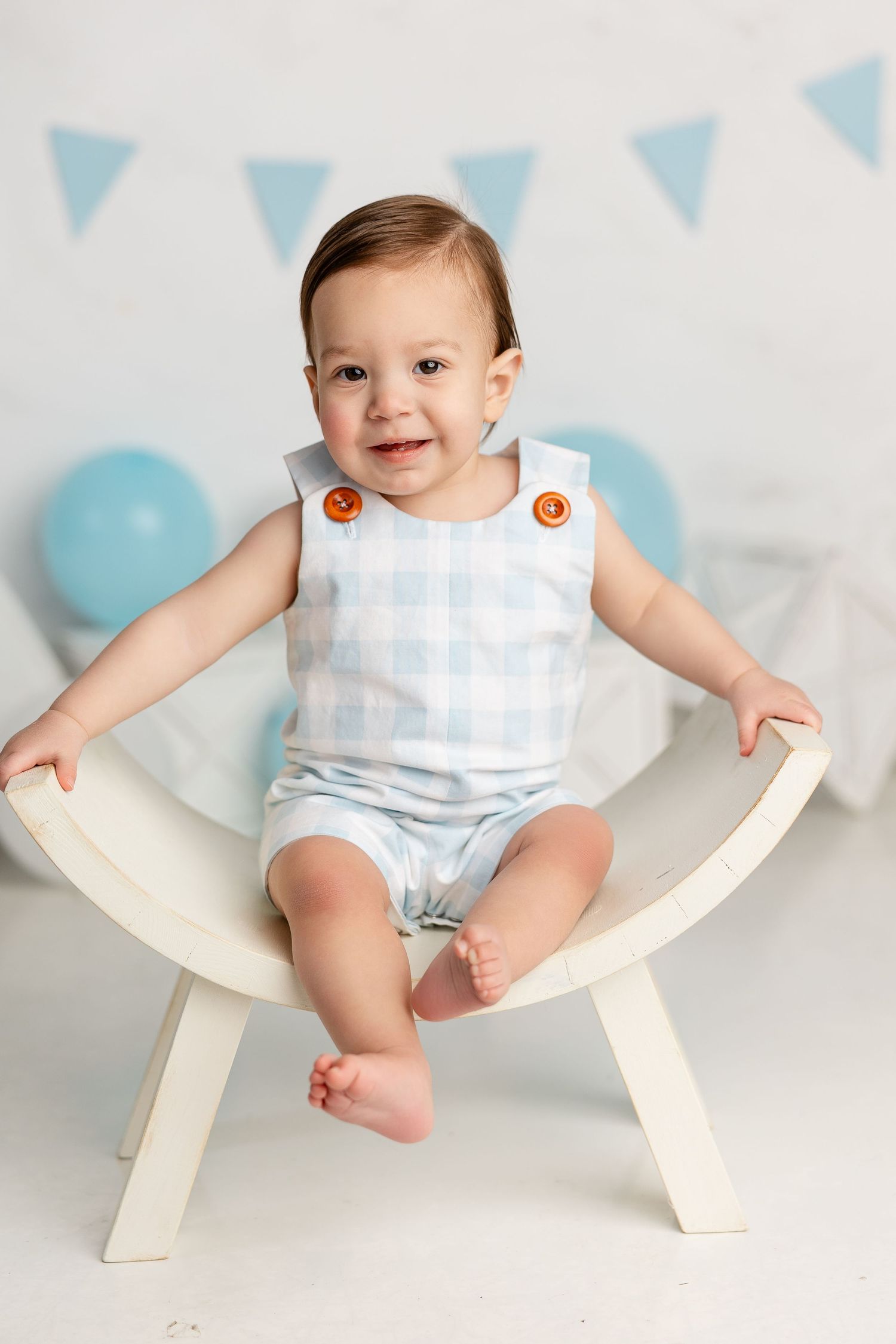 Baby in white and blue outfit sits smiling in curved wooden chair against backdrop with blue balloons and bunting.