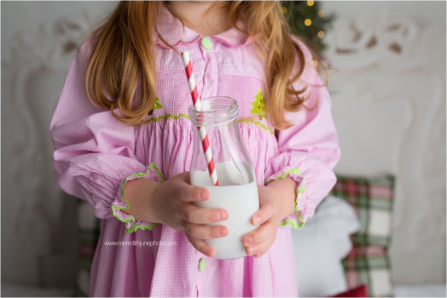 Child in pink nightgown holds glass of milk and cookies near decorated Christmas tree.