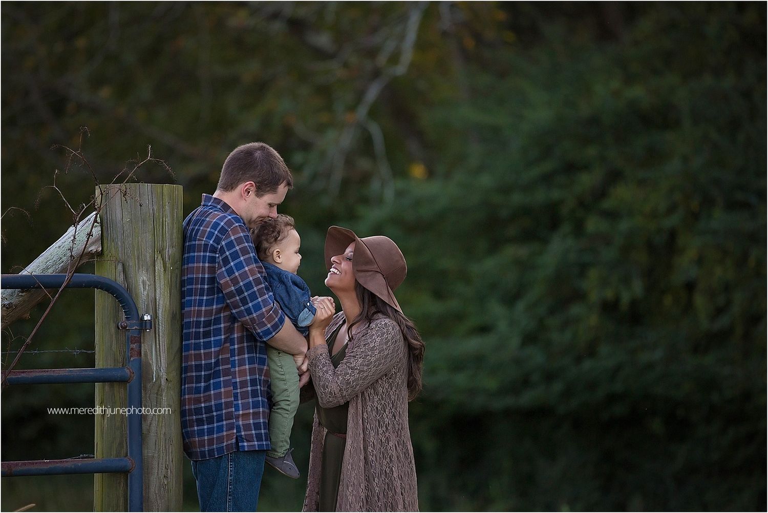 A couple shares an intimate moment by a wooden fence post at dusk with trees in the background.